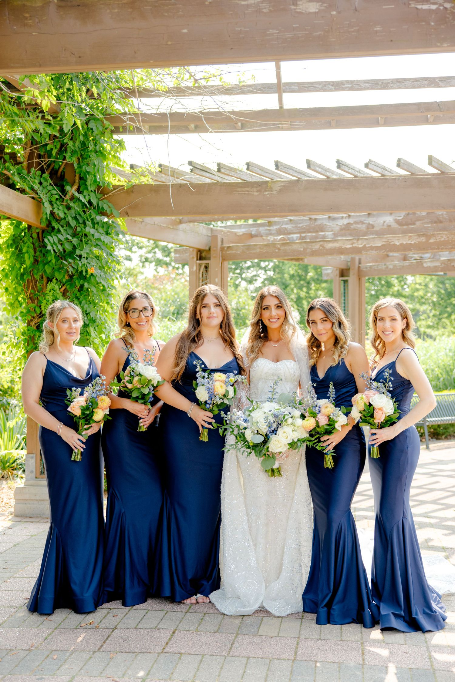 Wedding party in navy blue bridesmaid dresses poses with bride under rustic pergola.