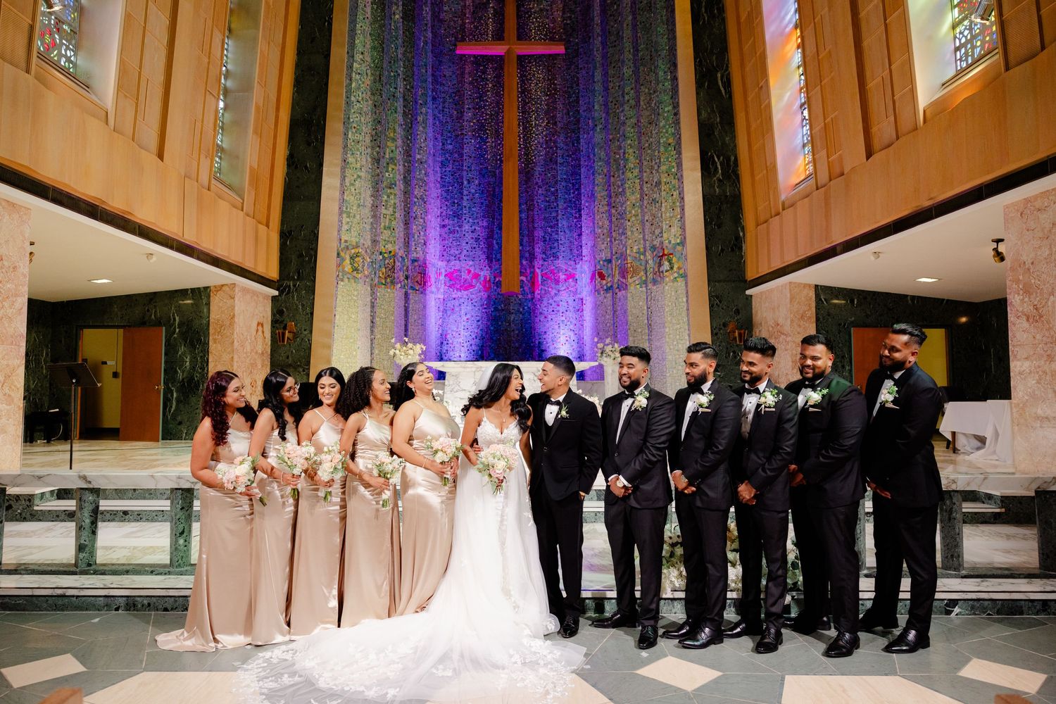 Wedding party with bridesmaids in beige and groomsmen in black pose at illuminated church altar.