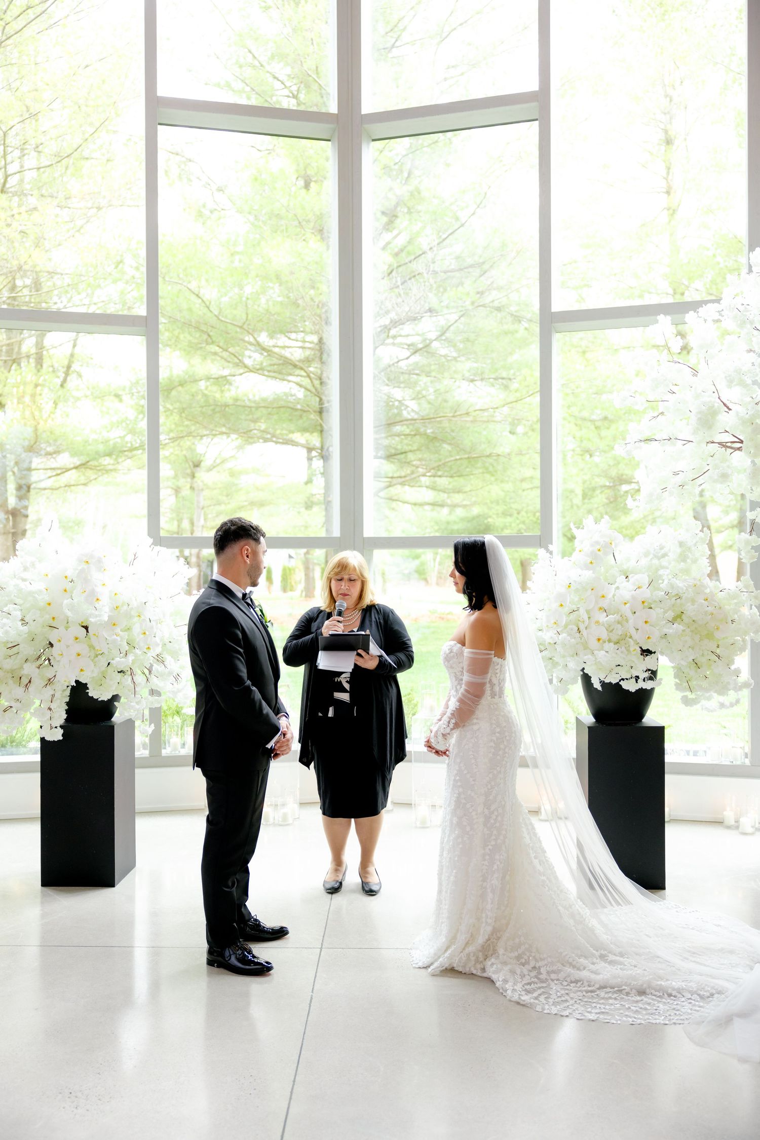 A wedding ceremony takes place in a bright modern venue with floor-to-ceiling windows and white floral arrangements.