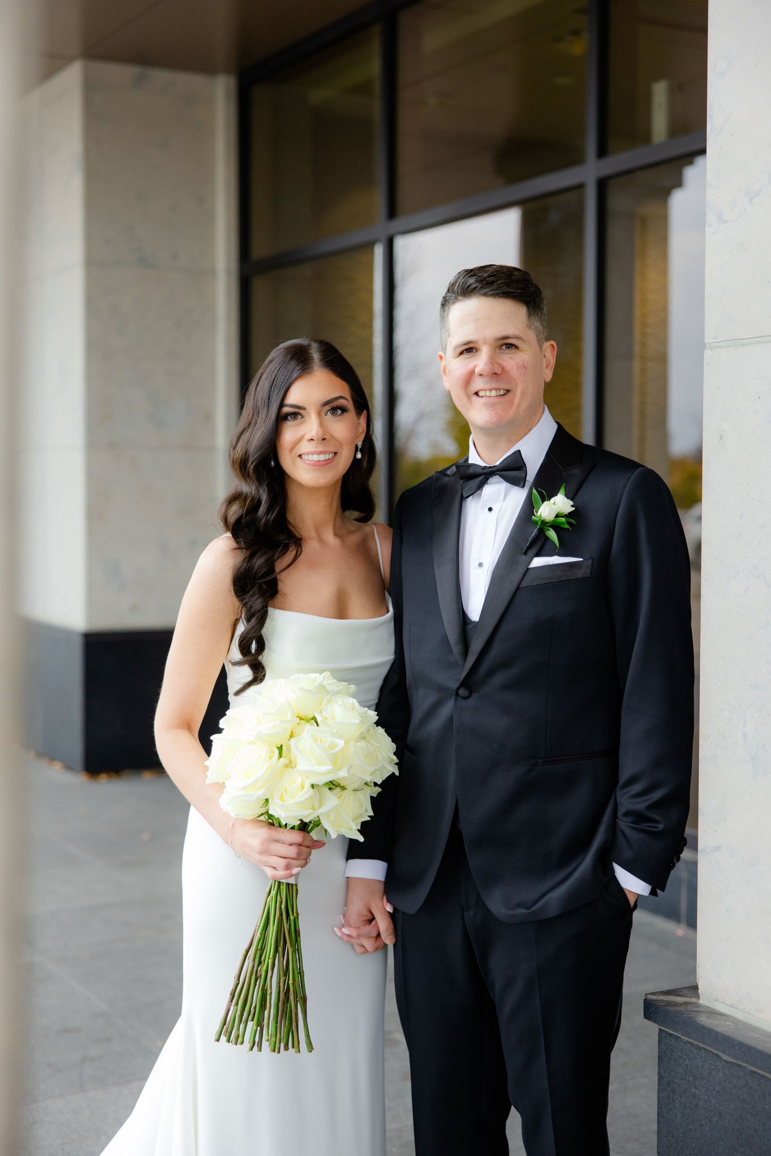 Elegant bride in white gown holds cream bouquet while posing with groom in classic black tuxedo outdoors.