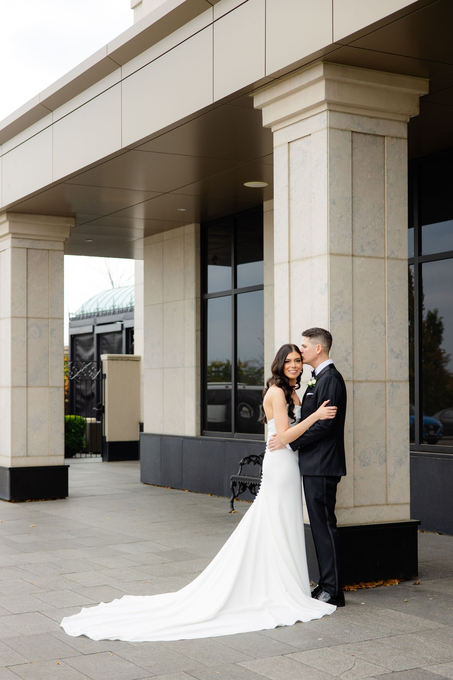 Romantic wedding portrait of couple embracing under modern building colonnade.