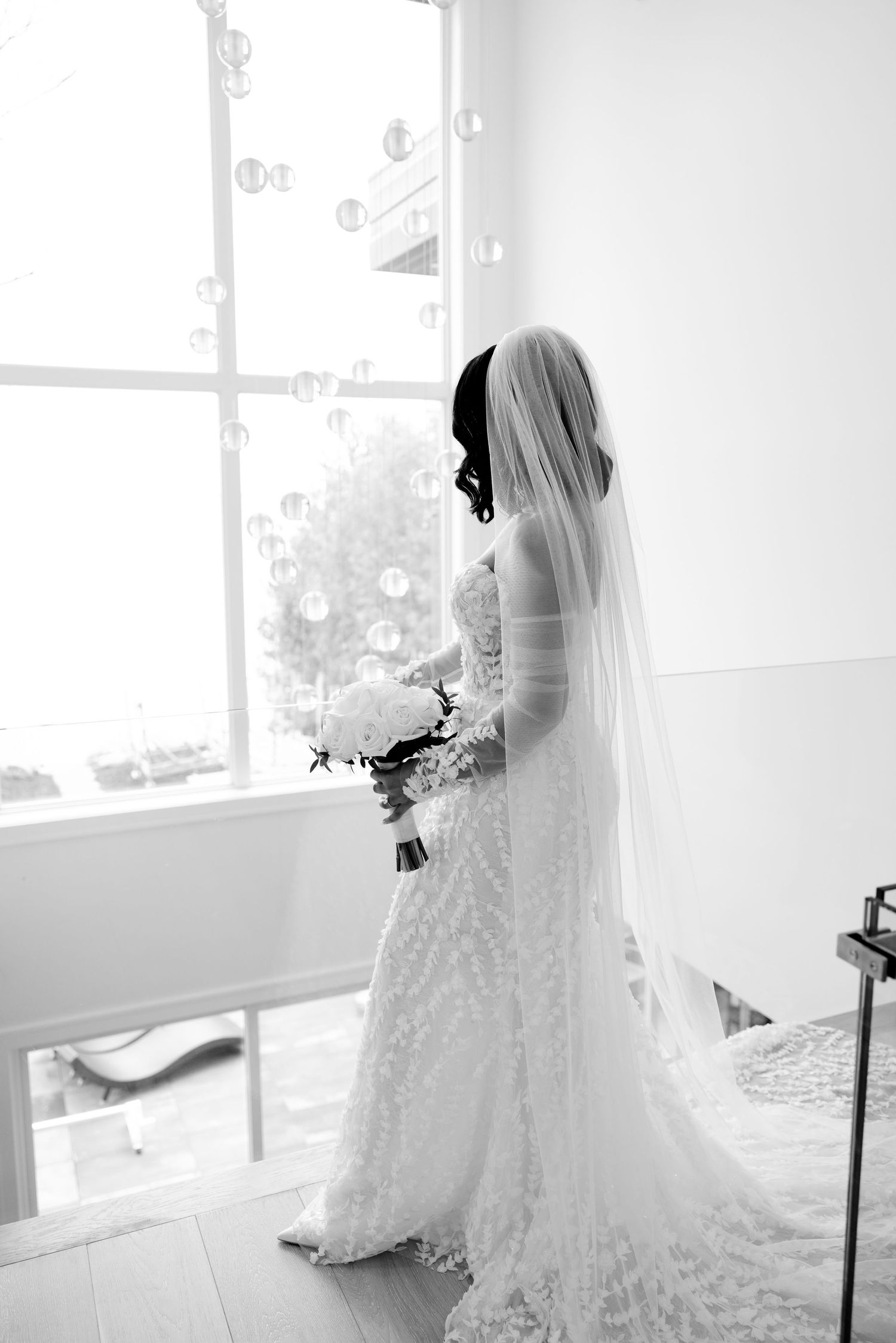 A bride in an elegant lace wedding gown and veil stands by a bright window holding a bouquet.