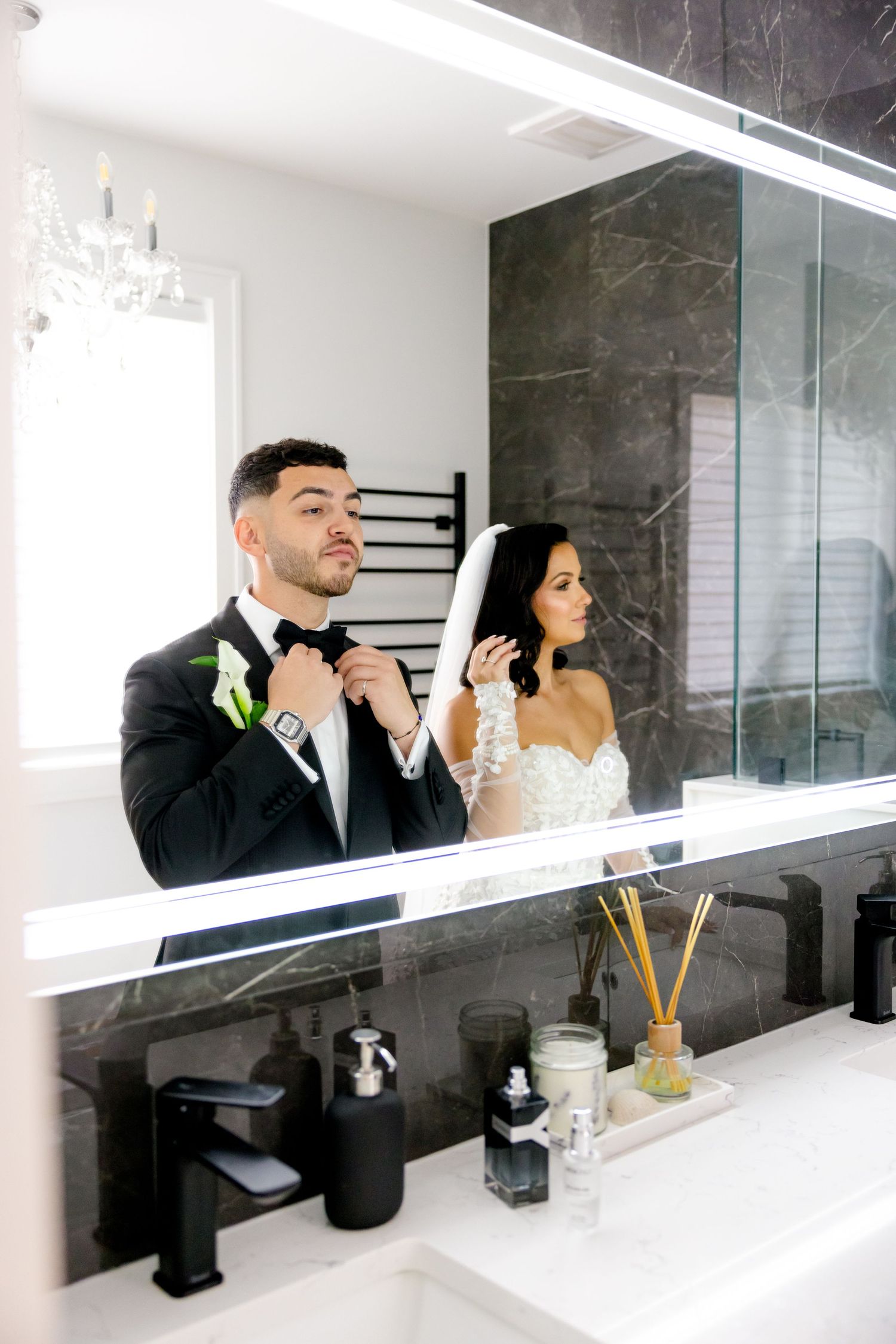 Reflection of a couple in formal wedding attire adjusting their accessories in a bathroom mirror.