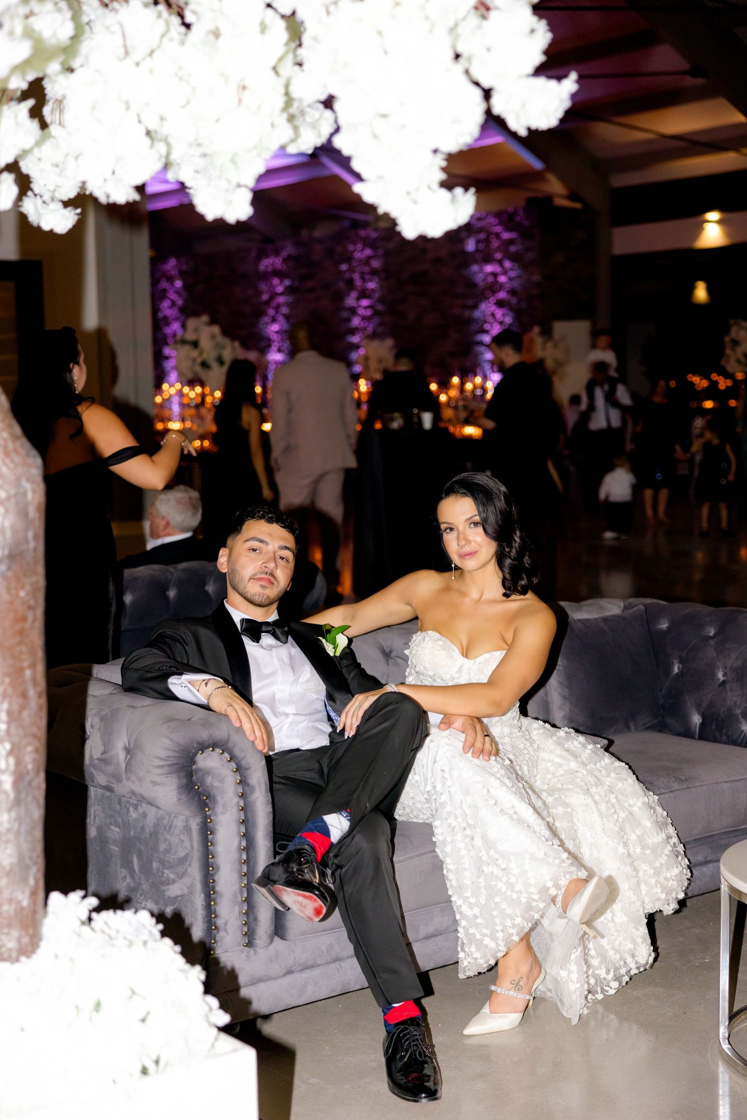Couple relaxing on gray couch at wedding reception under purple lighting and white floral decor.