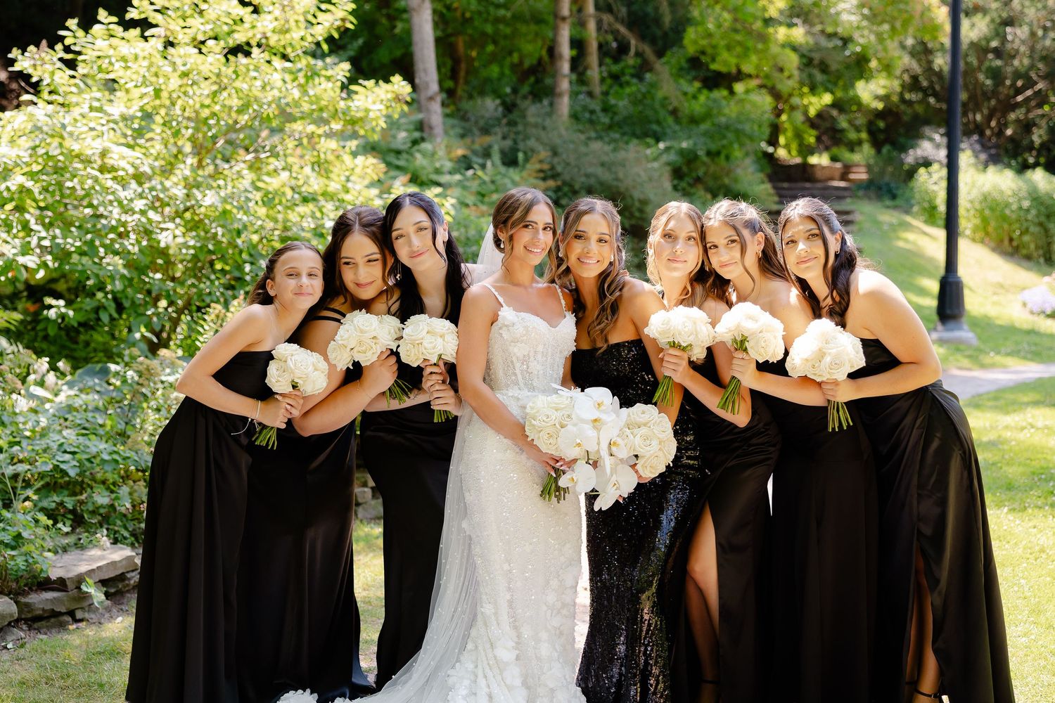 A wedding party poses for portraits in a sunny outdoor garden setting wearing black and white formal attire.