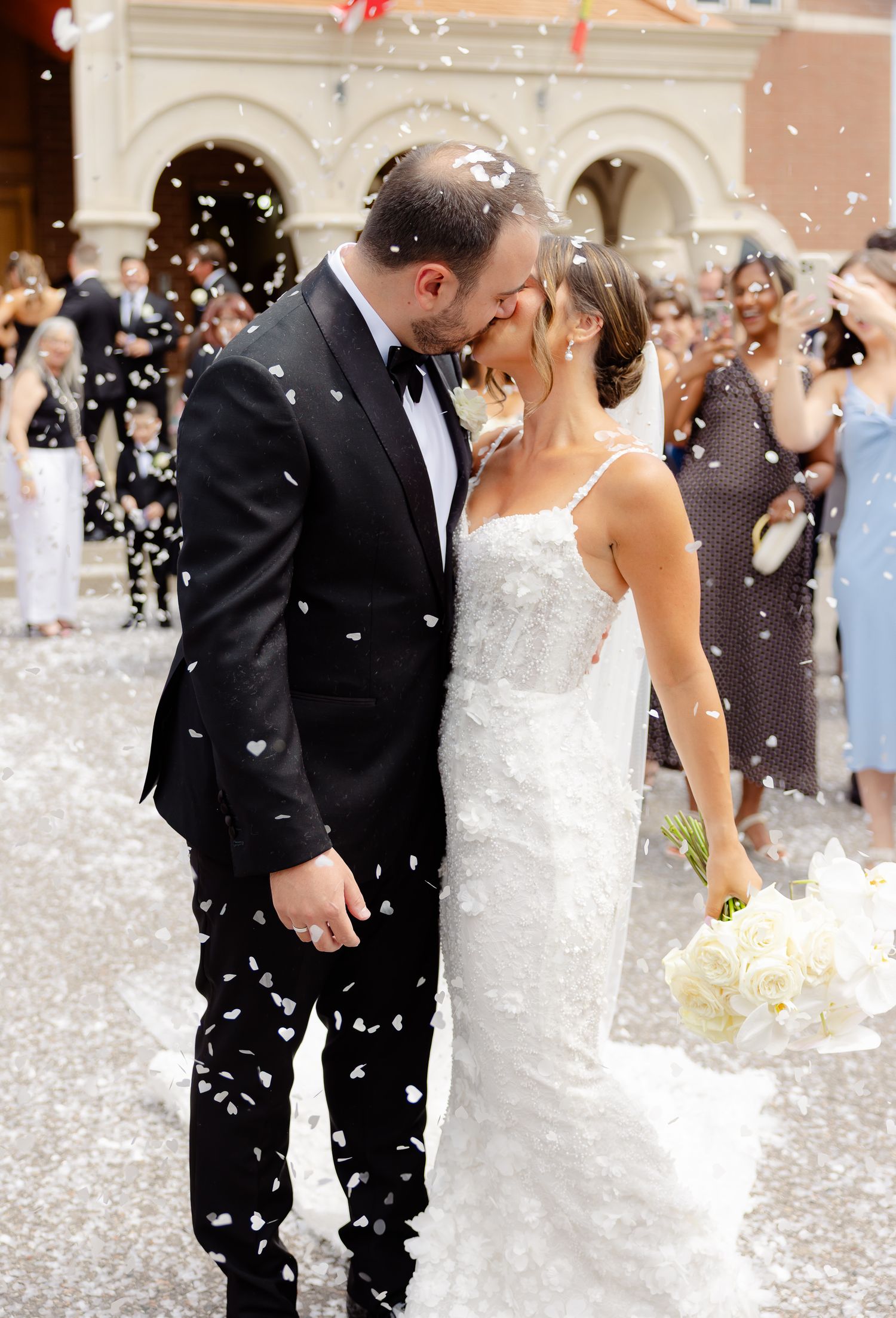 A wedding couple shares an intimate moment under a shower of confetti in front of an elegant arched entrance.