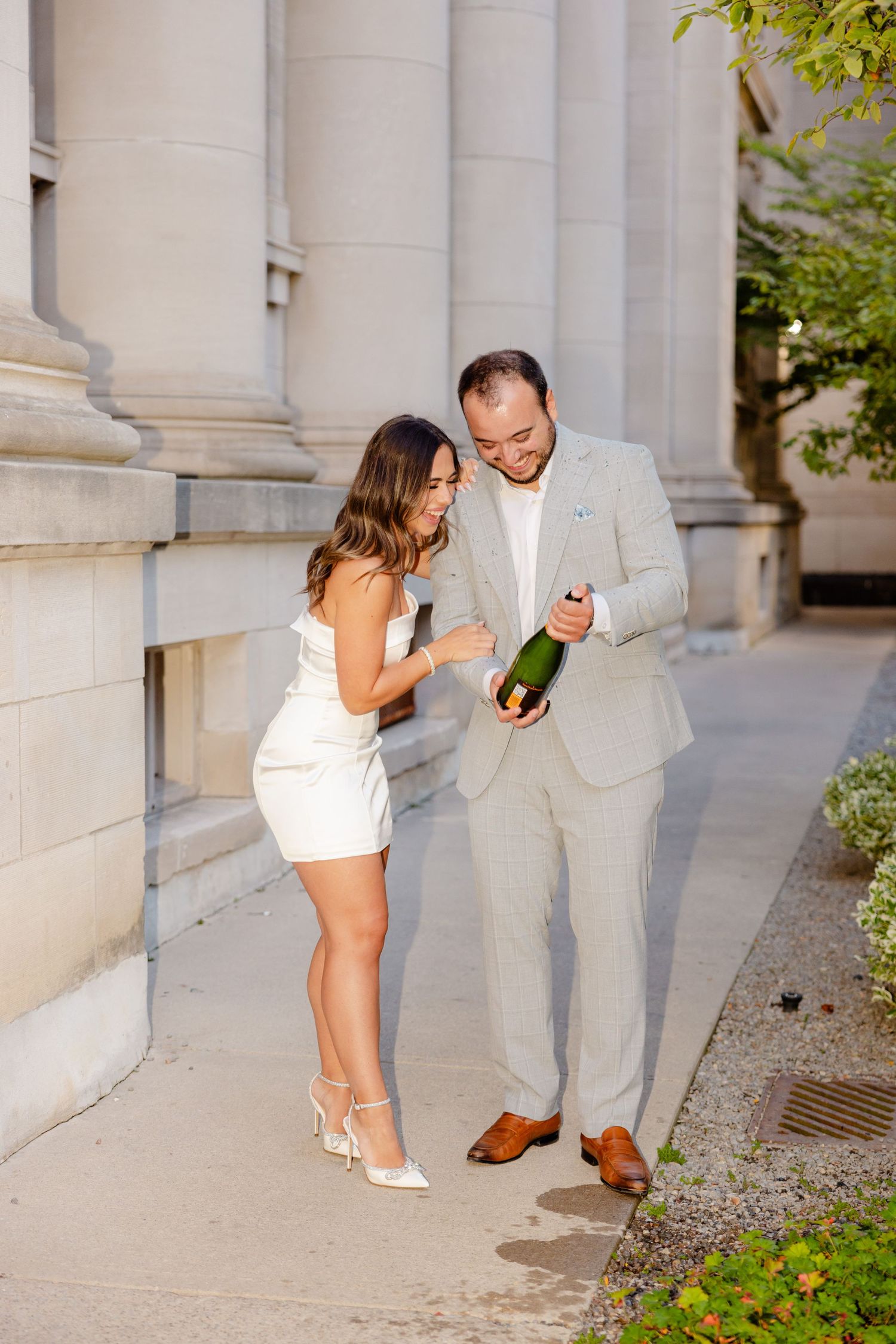 A couple shares an animated moment while handling champagne near classical architecture.