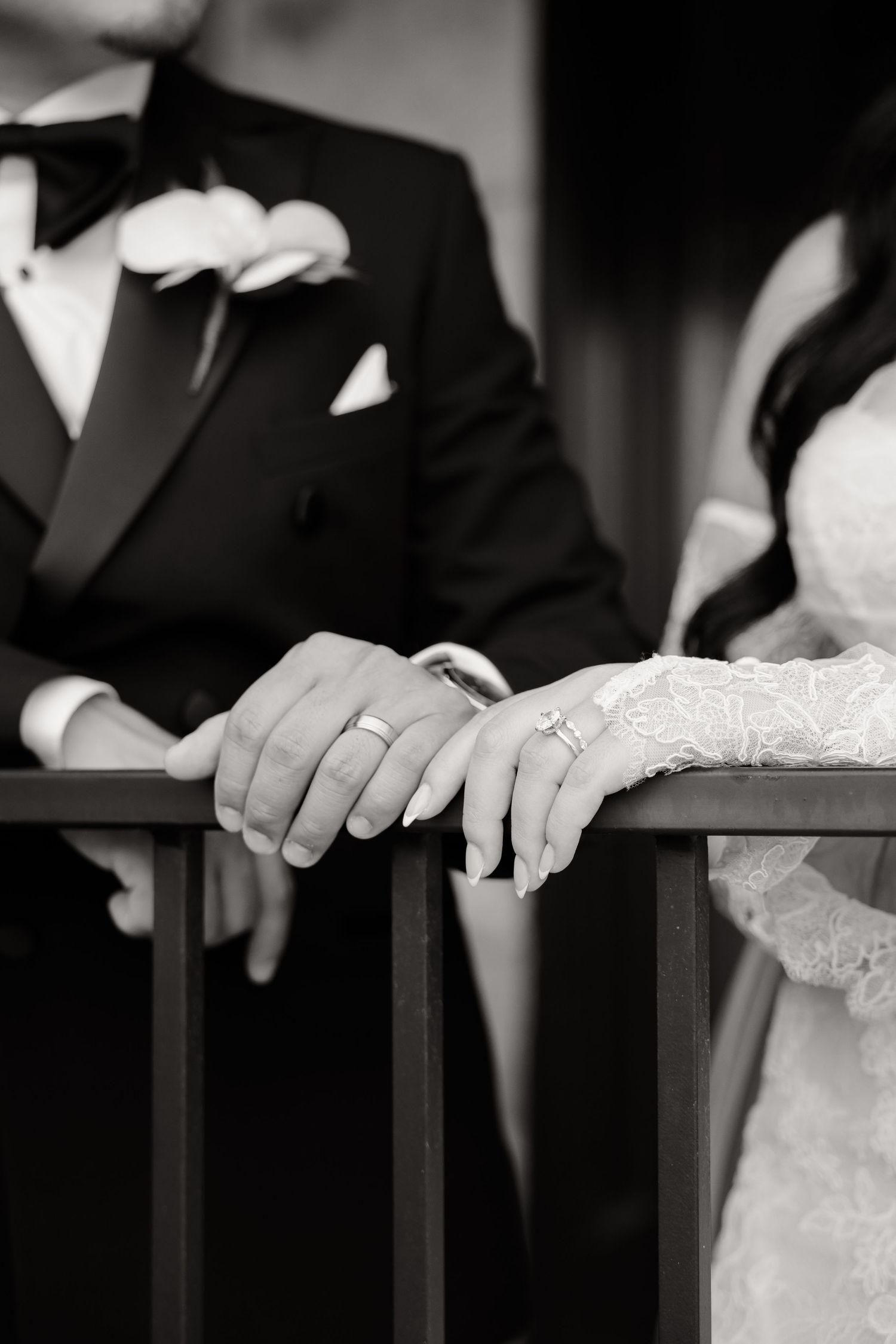 A black and white close-up photograph showing hands resting on a railing in formal wedding attire.