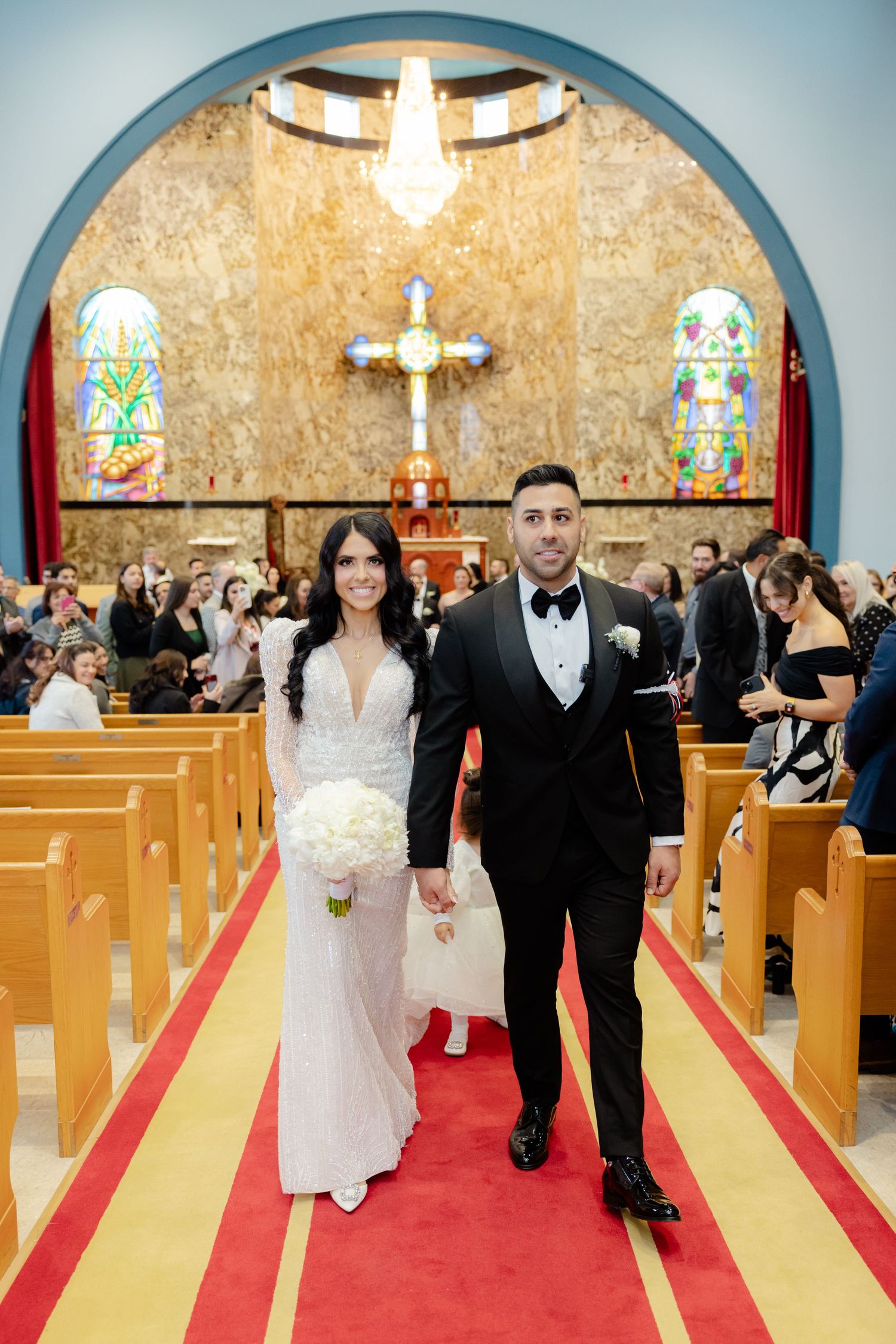 Elegant couple walks down church aisle with ornate cross and stained glass windows behind them.