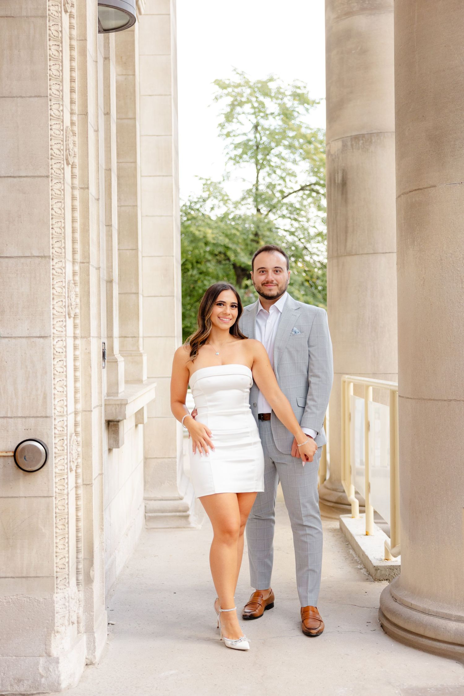 A couple poses elegantly between stone columns wearing a white dress and light gray suit.