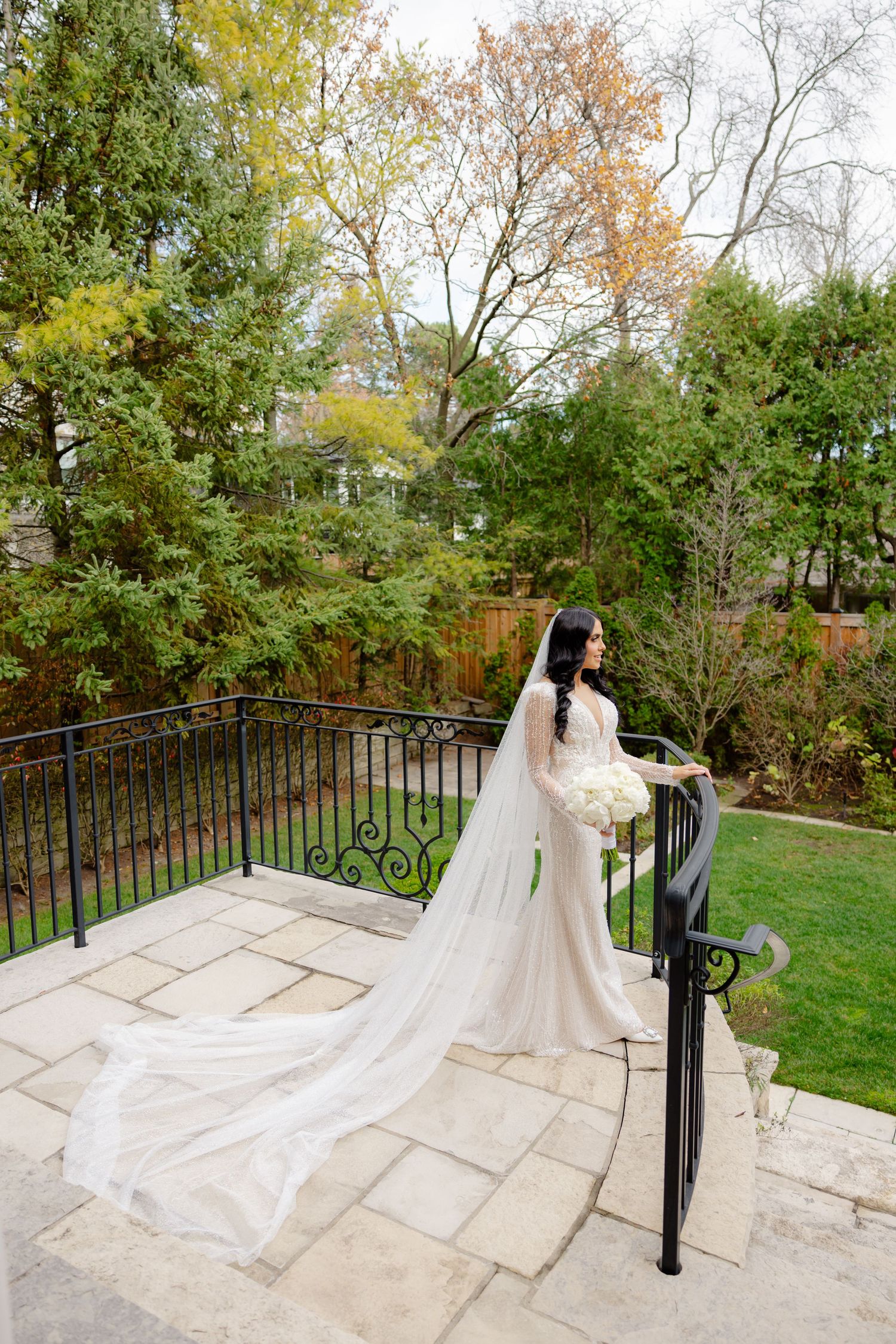 Bride with flowing cathedral veil stands at wrought iron railing overlooking autumn garden.