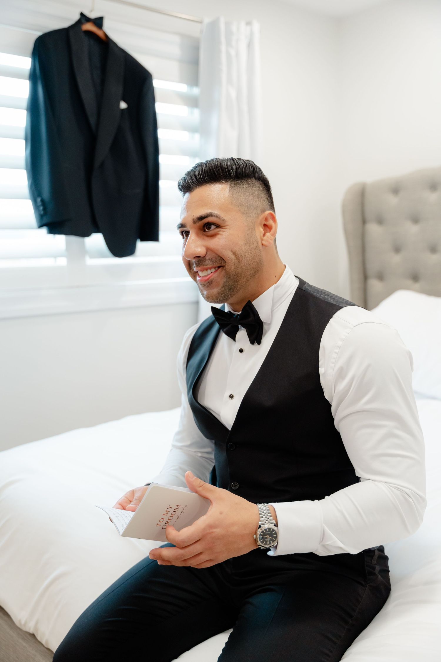 Person in formal vest smiling while holding a note, with suit jacket hanging in background.