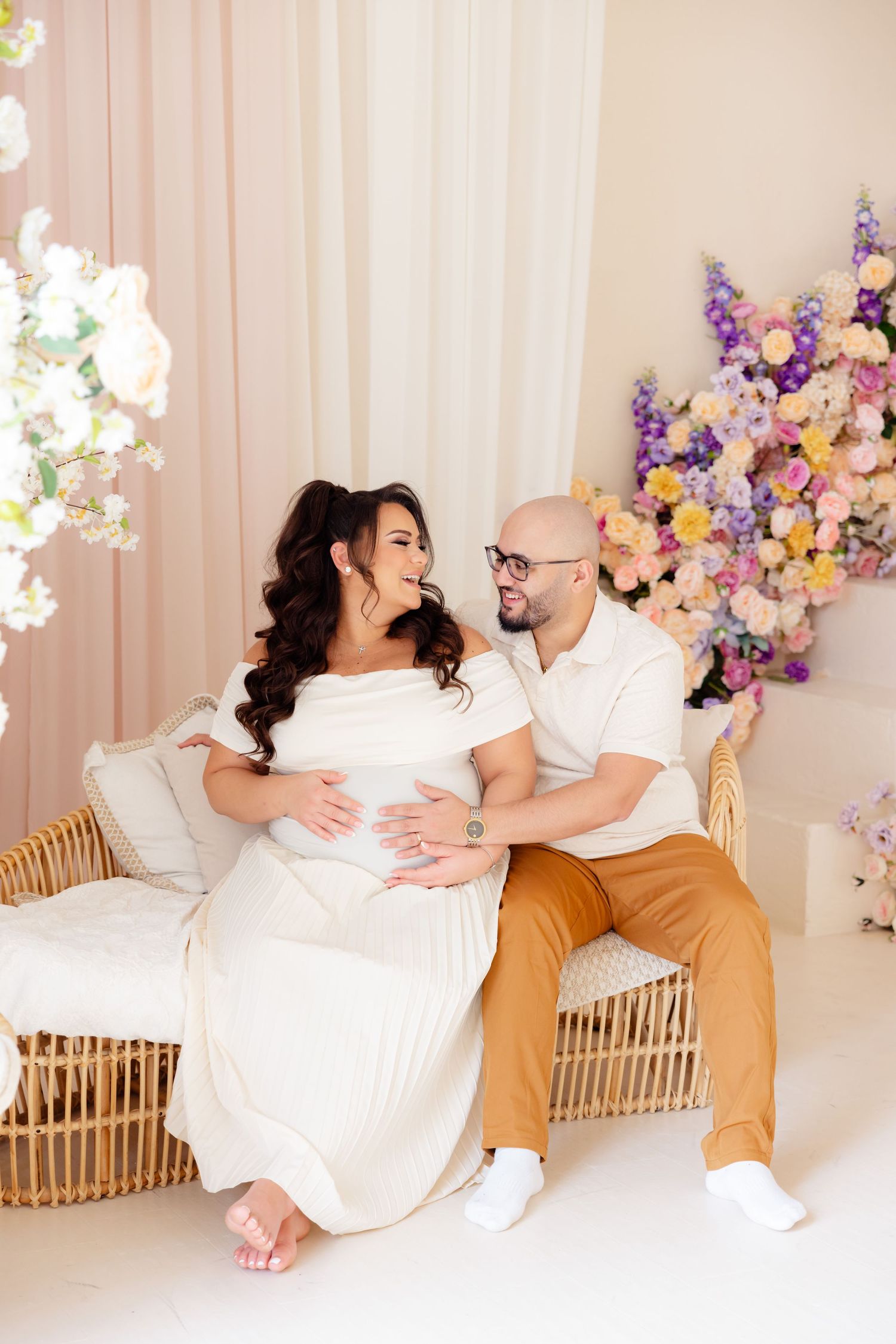 A couple sits together on a rattan bench with colorful floral arrangements decorating the background setting.