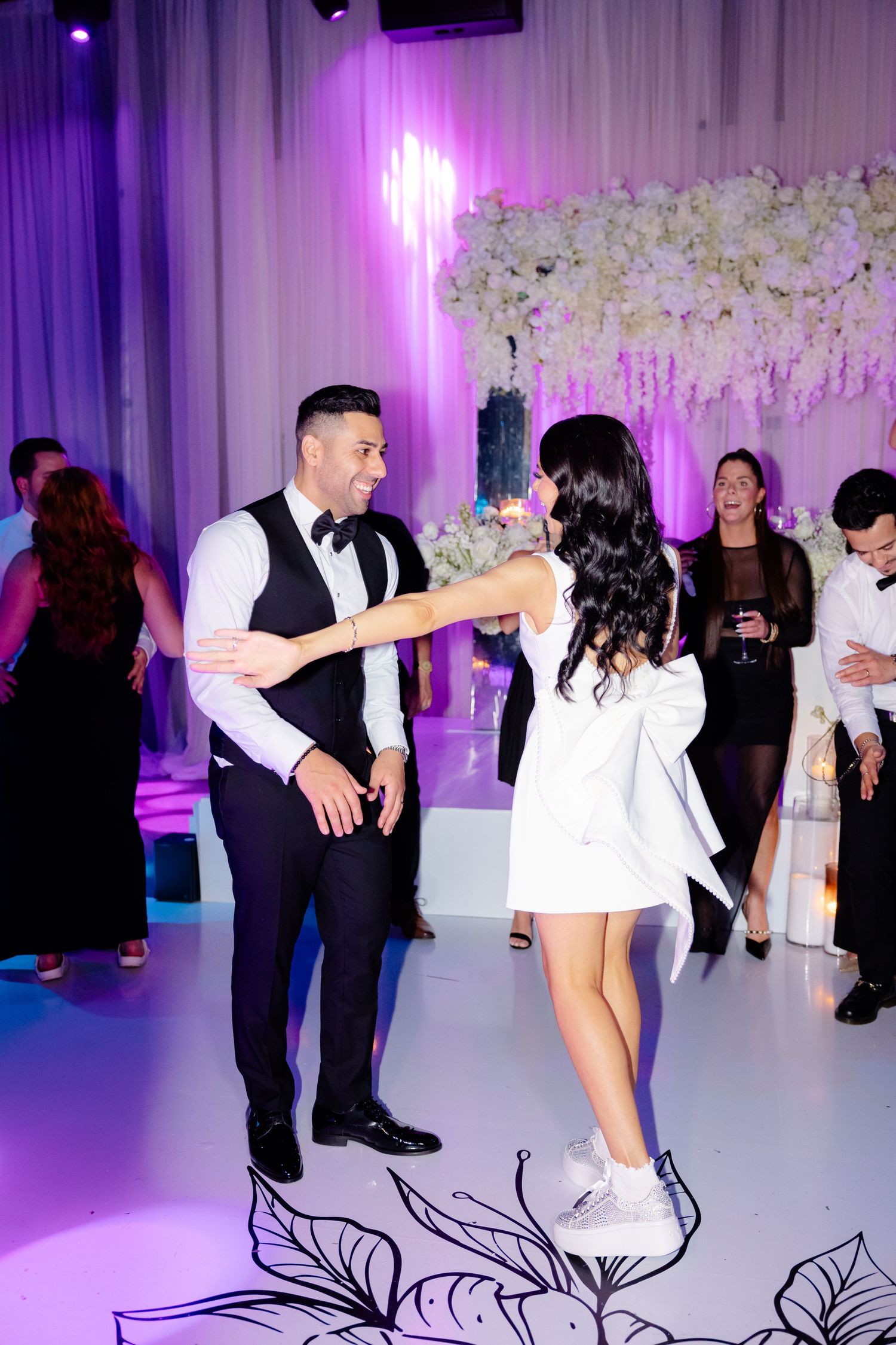 Couple dancing in formal attire at an elegant wedding reception with purple lighting and floral decor.