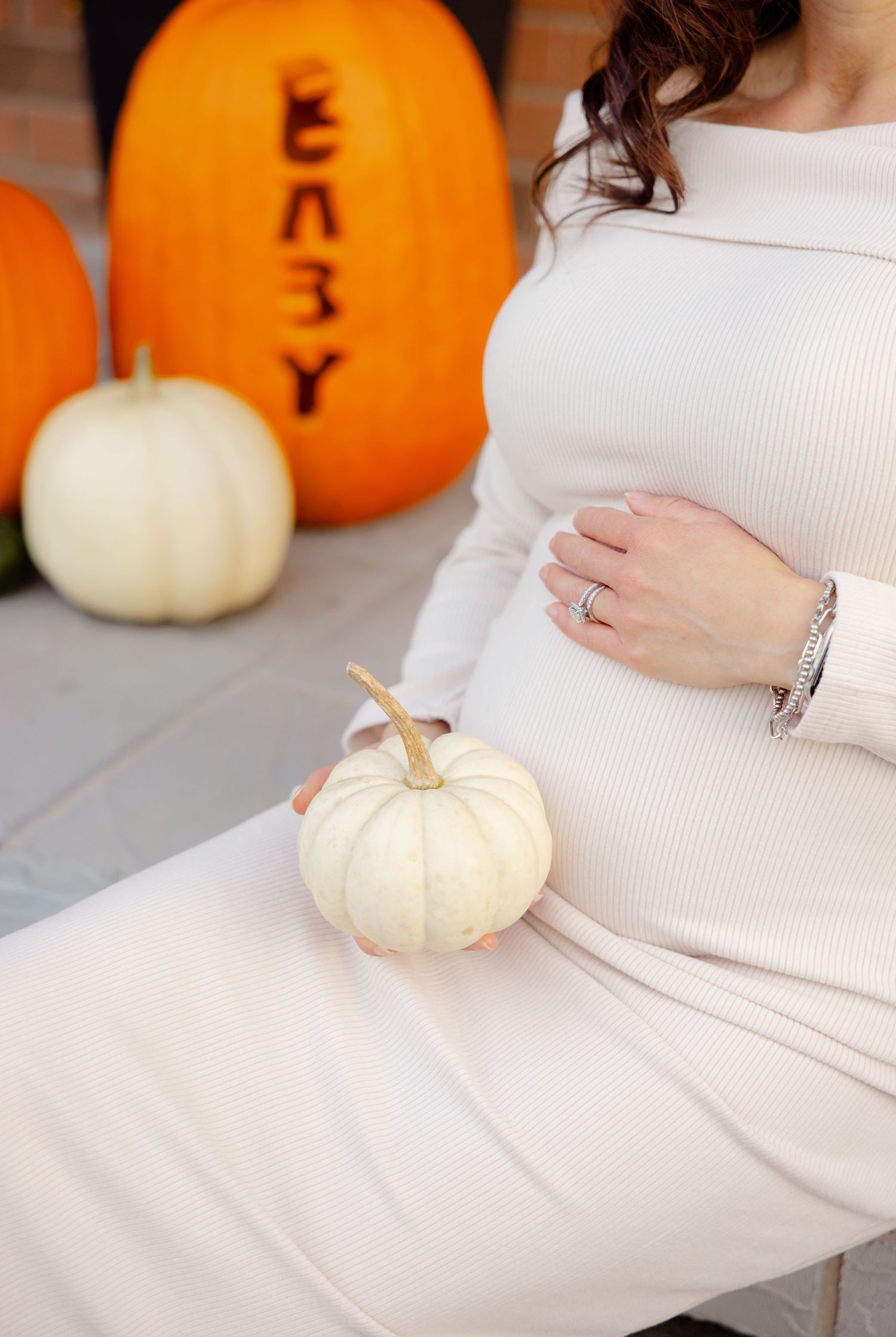 Pregnant woman in white dress sits with white pumpkin, orange pumpkin with 'BABY' text in background.