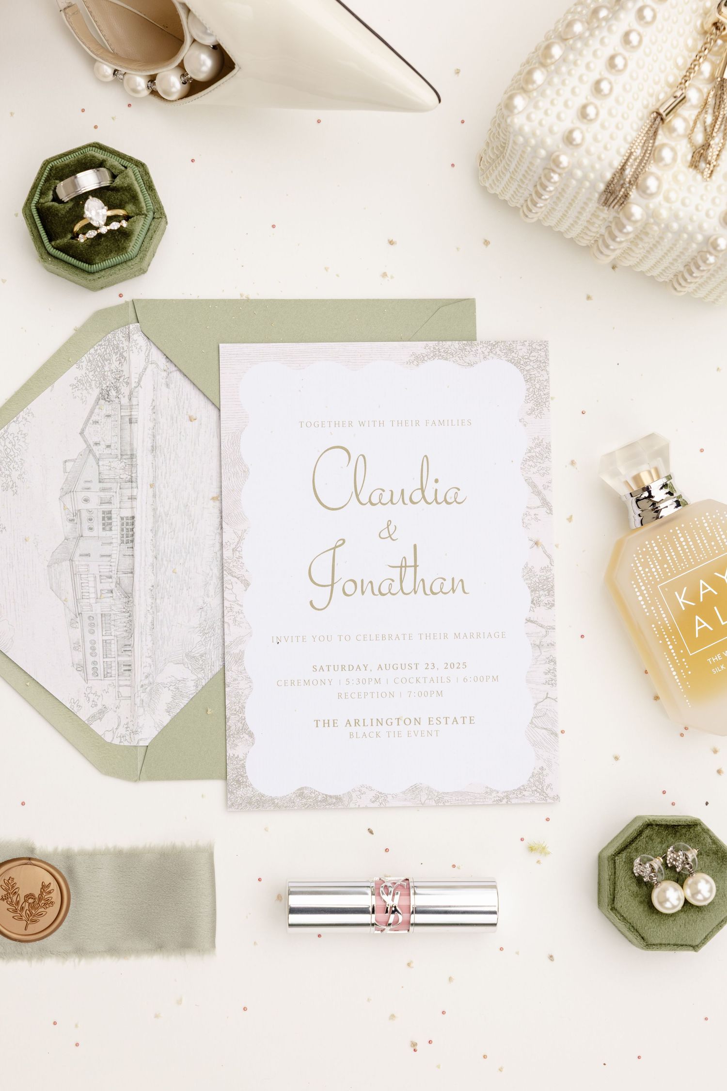 Classic wedding invitation suite with sage green envelope, perfume bottle, and pearl accents on white surface.