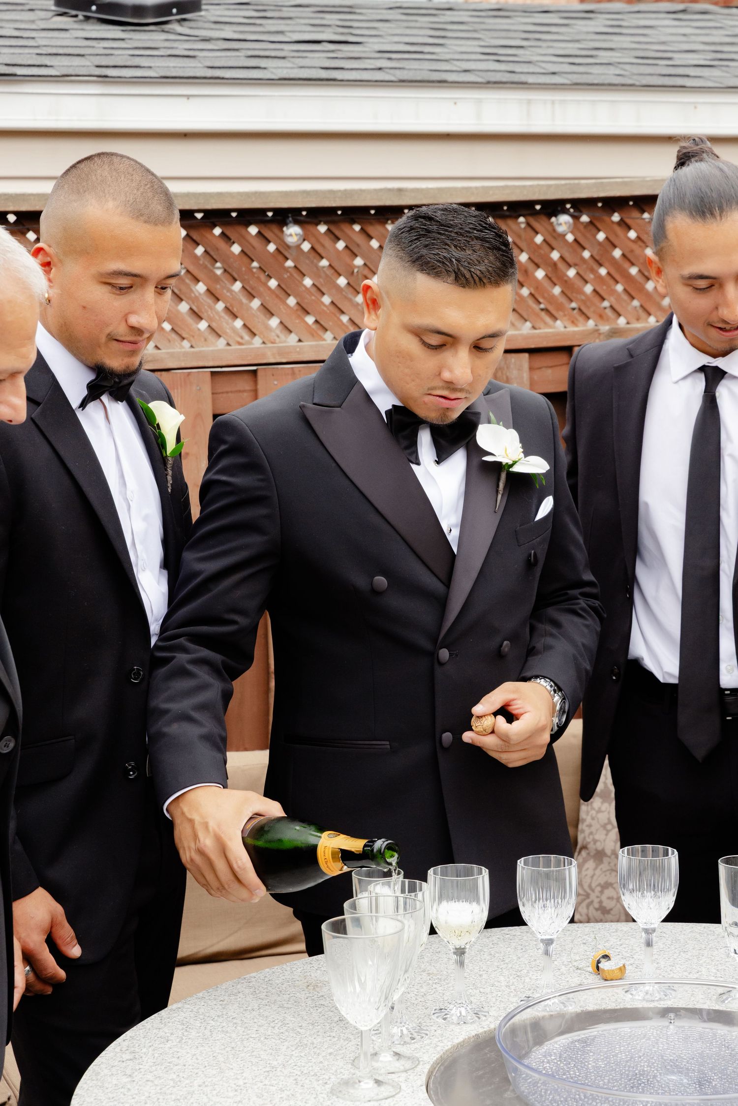 Groomsmen in black suits and ties pour drinks at an outdoor event bar.