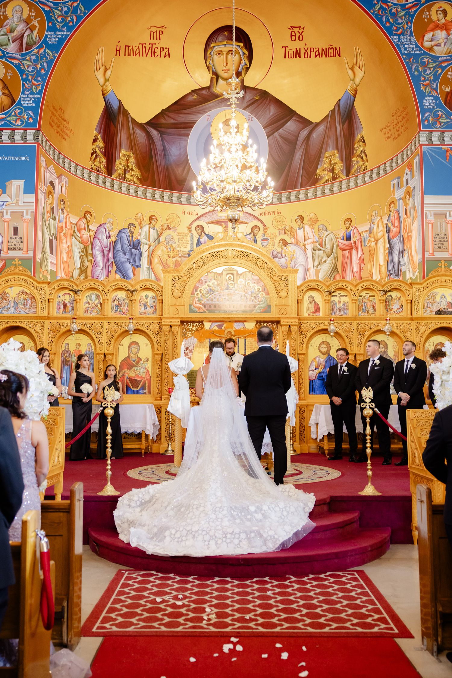 An Orthodox Christian wedding ceremony takes place inside an ornate church with golden icons and chandeliers.
