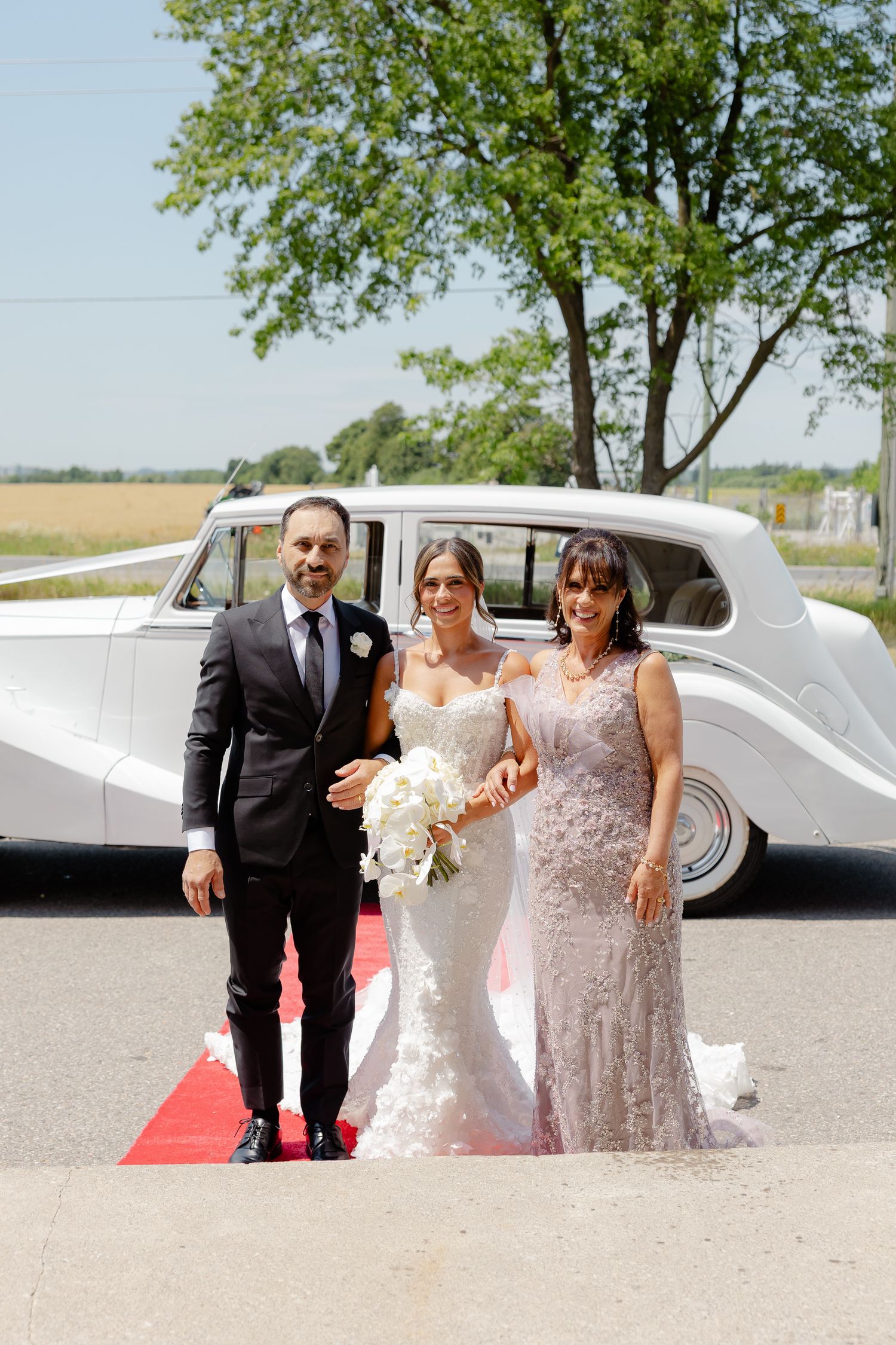 An elegant wedding party poses by a vintage white Rolls Royce on a red carpet in a rural outdoor setting.