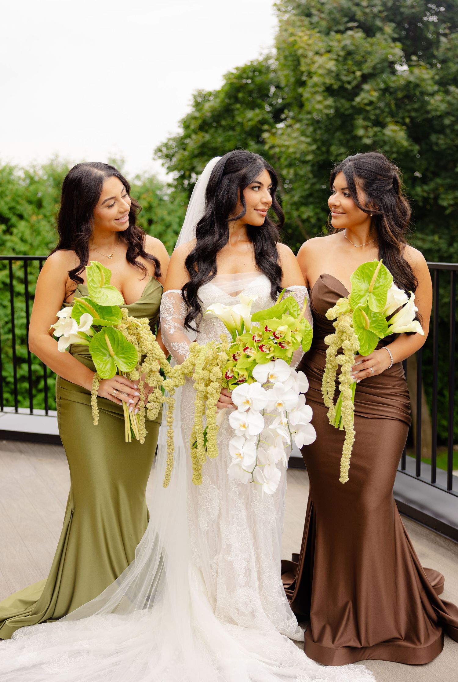 Bridesmaids in olive green and brown satin dresses stand together holding white orchid bouquets during an outdoor wedding.