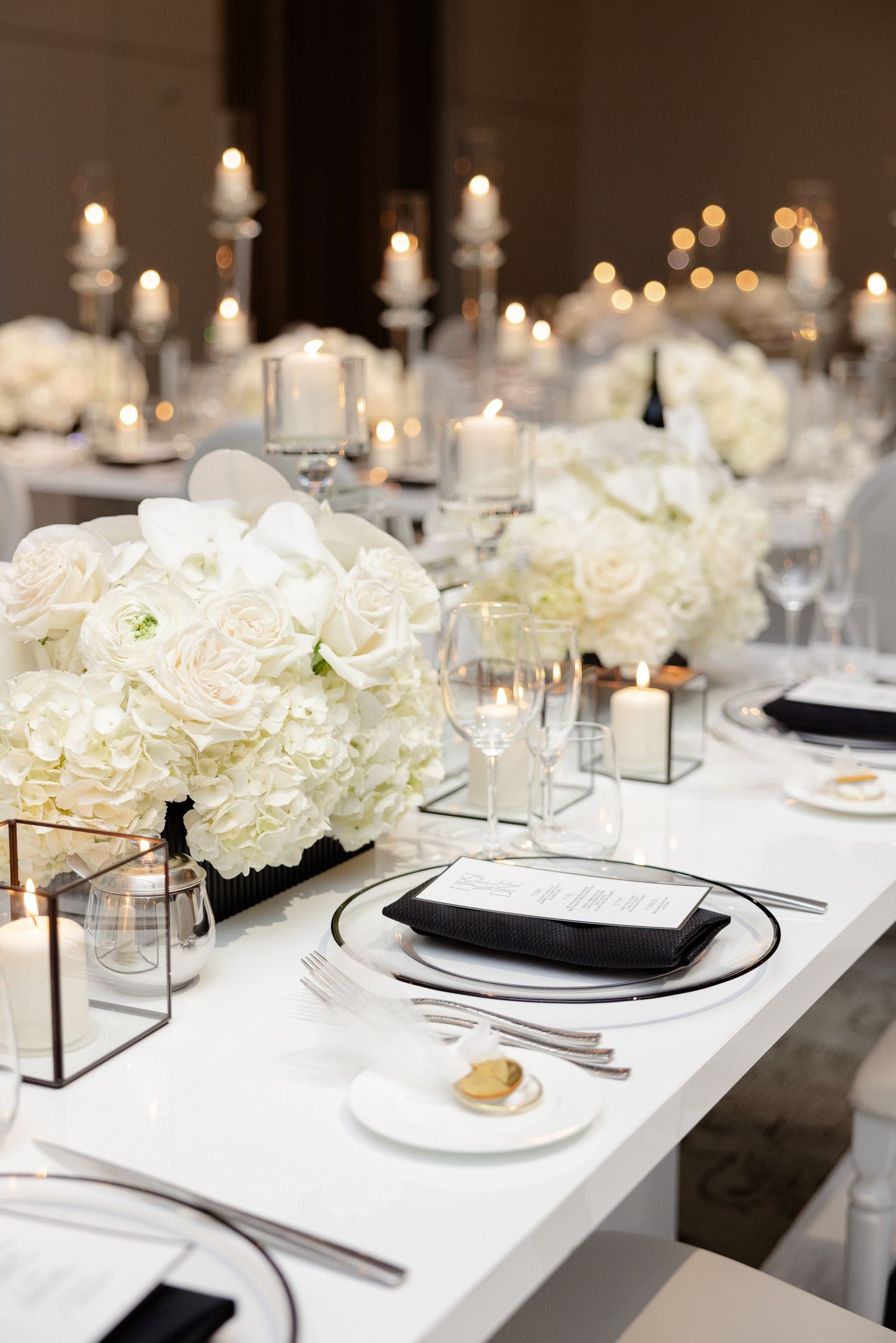 Elegant white wedding table setting with hydrangea centerpieces, candles, and modern black and white place settings.