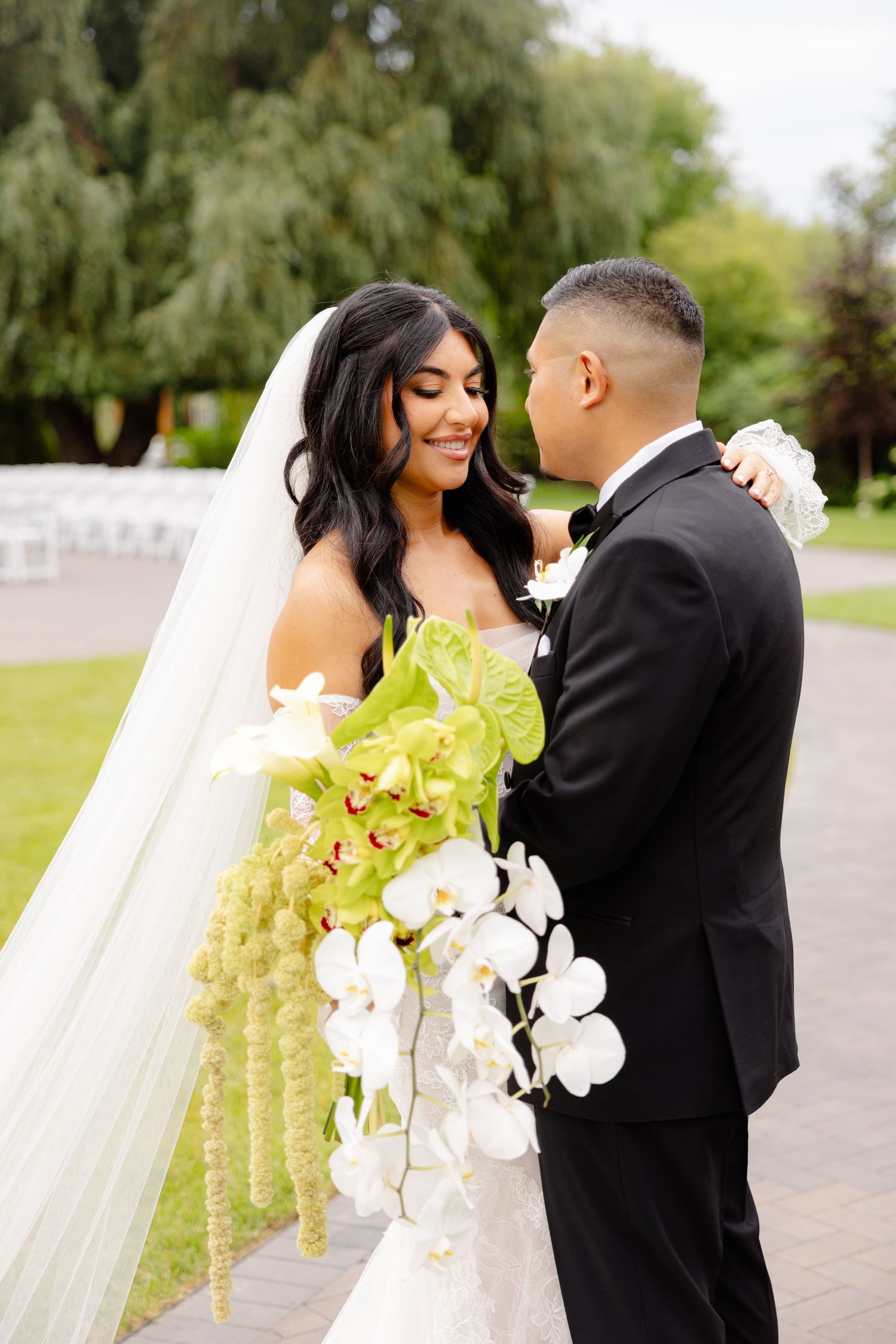 A tender embrace between a wedding couple outdoors with a stunning cascading bouquet of white orchids and green flowers.