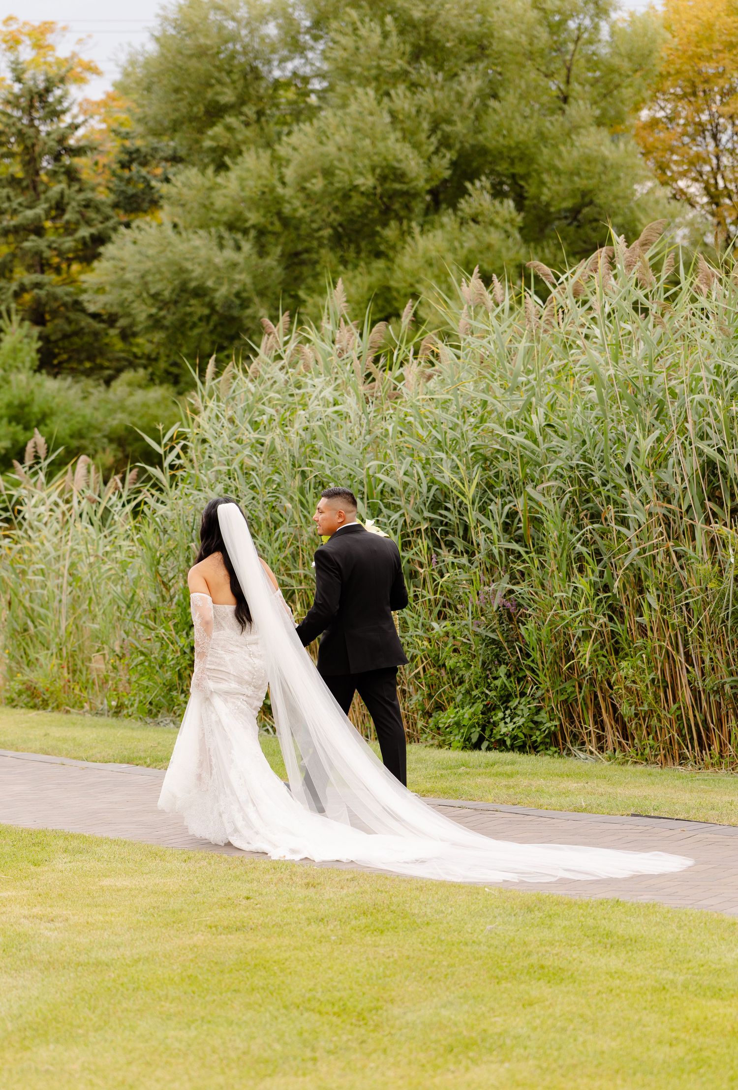 A couple in wedding attire stand on a path surrounded by tall golden grasses and autumn foliage.