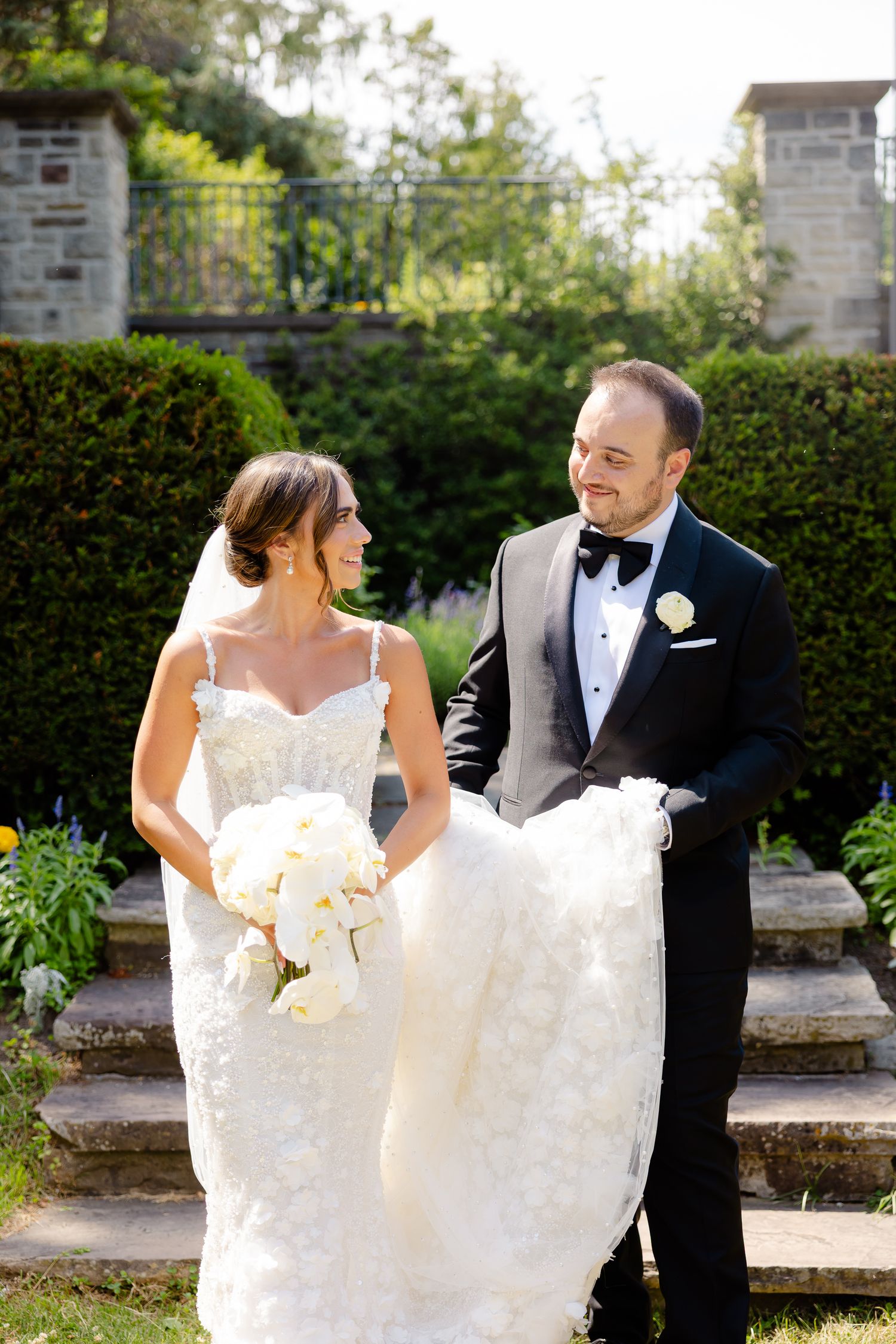A romantic newlywed couple gazes at each other while walking through an elegant garden setting during their outdoor wedding.