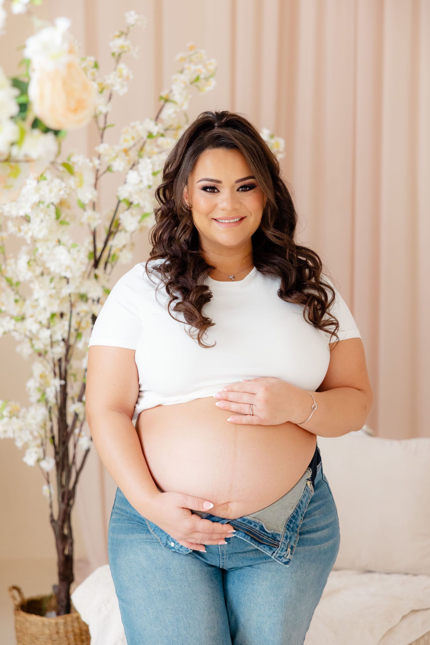 A smiling pregnant woman in a white t-shirt and jeans poses with her hands on her belly in front of white floral decor.