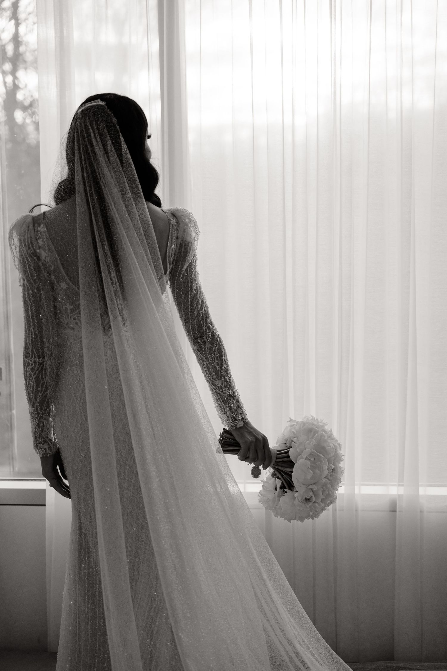 Black and white photo of bride with flowing veil holding white bouquet by window.