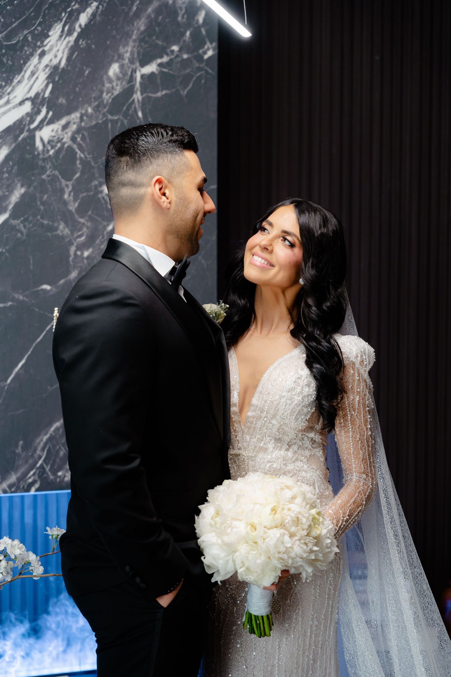 Newlyweds share an intimate moment against dark marble wall with blue decor accents.