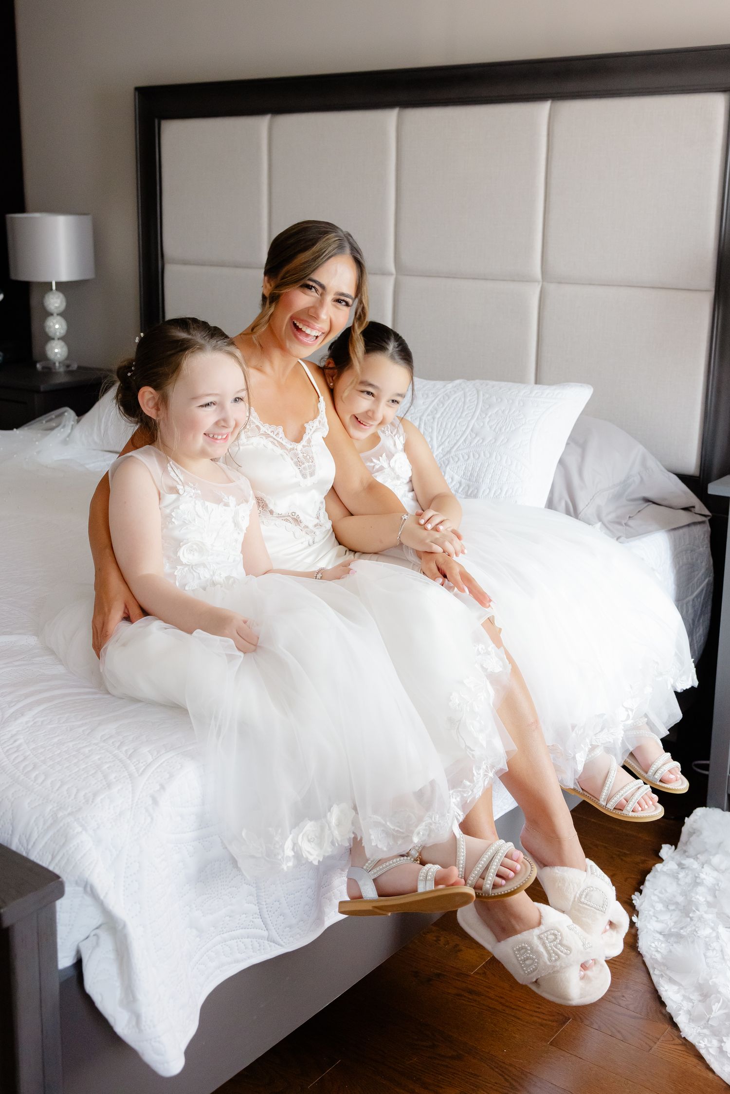 Three people in matching white dresses sit together on a bed, sharing joyful smiles in a bright bedroom setting.