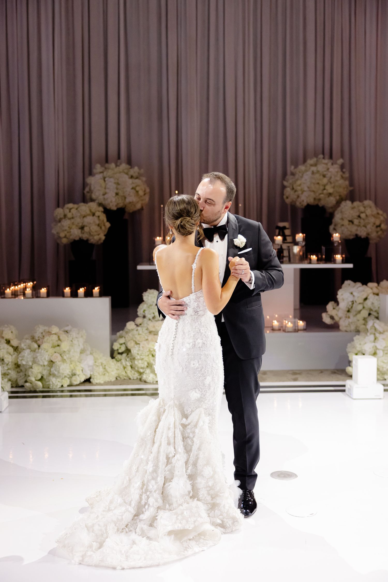 Two people share their first dance at an elegant wedding reception with white floral arrangements and candlelight decor.
