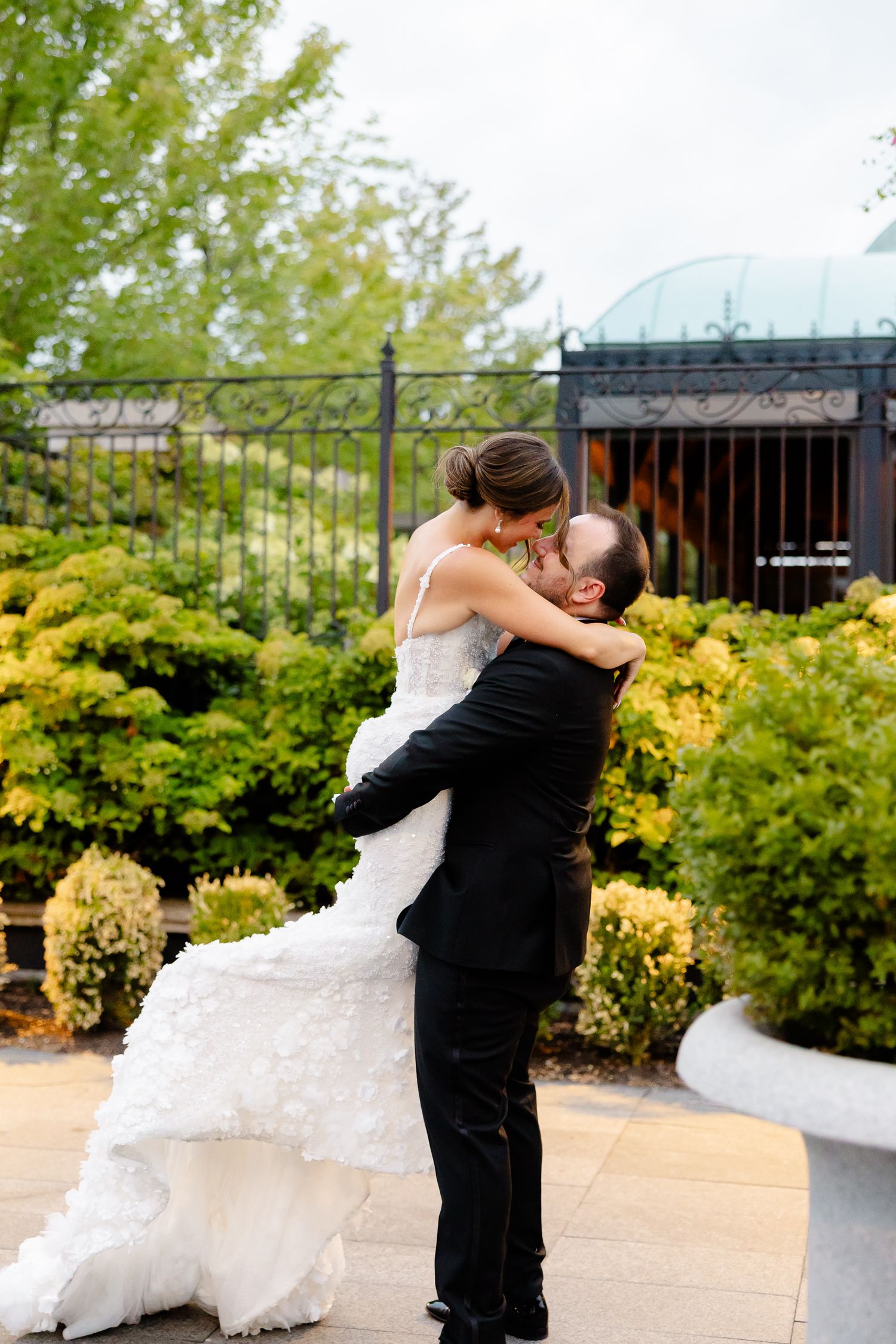 A bride and groom embrace in a romantic moment outdoors with lush yellow gardens and greenery in the background.
