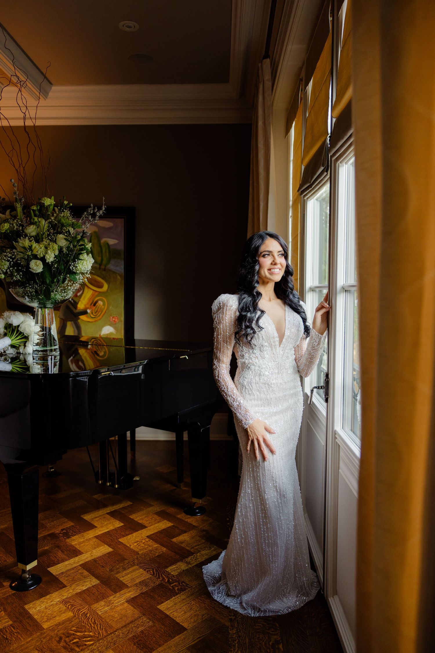 Wedding photo featuring a bride in white by a window with a grand piano and floral arrangement visible.