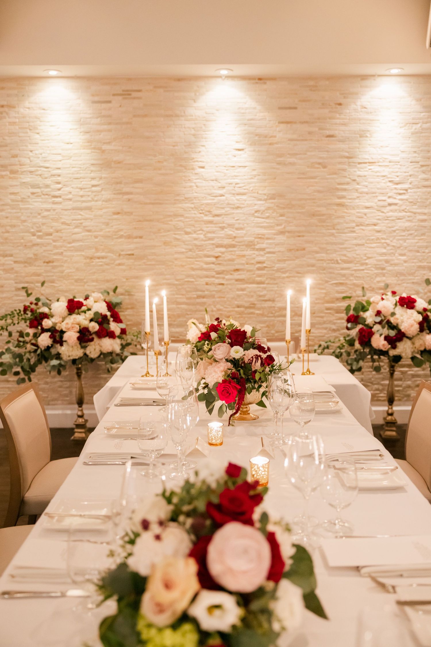 Long dining table with white linens adorned with red and white floral arrangements and tall taper candles.