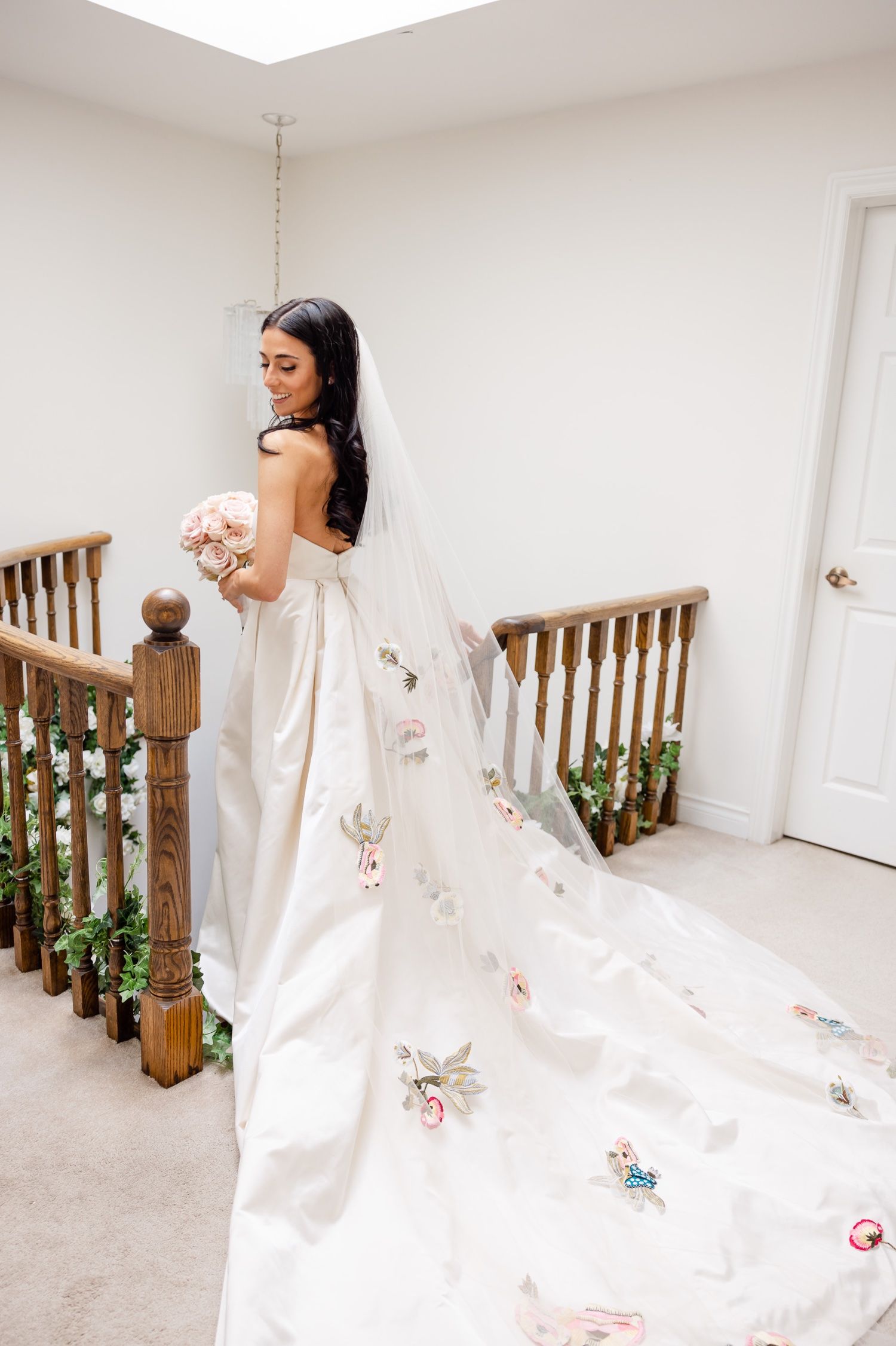 Bride in elegant white dress with floral decorated train poses on staircase.