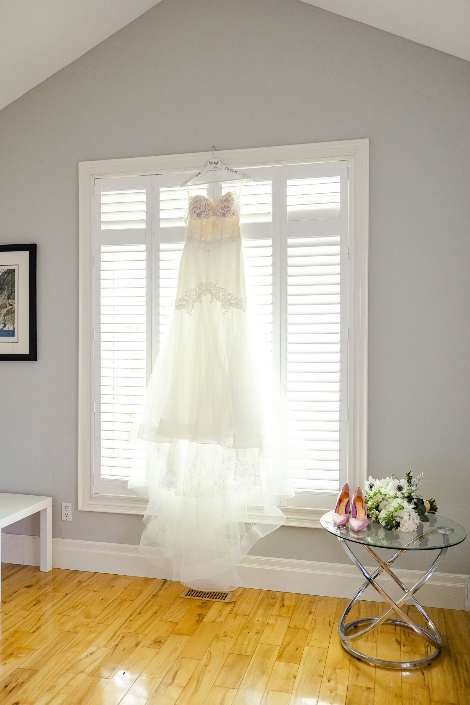 A delicate white wedding dress hangs in front of shuttered windows with a small side table displaying flowers.