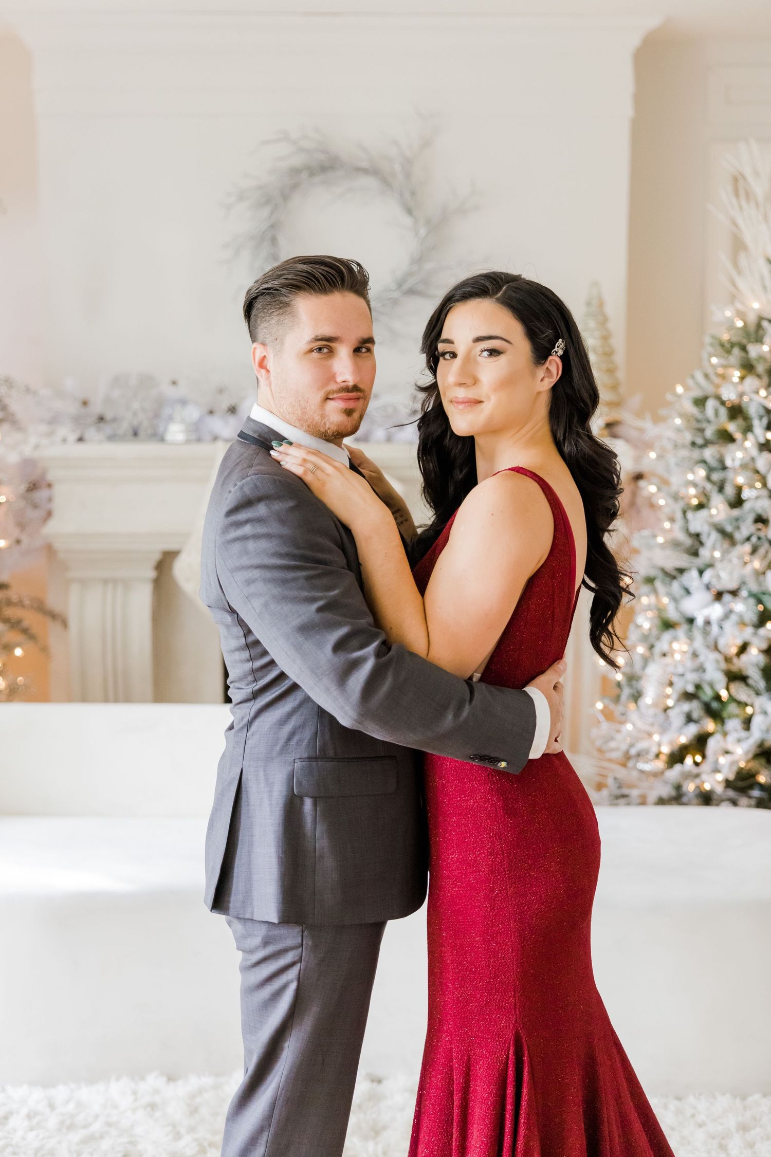 A couple poses elegantly in a winter wonderland setting with decorated Christmas trees and a red gown.