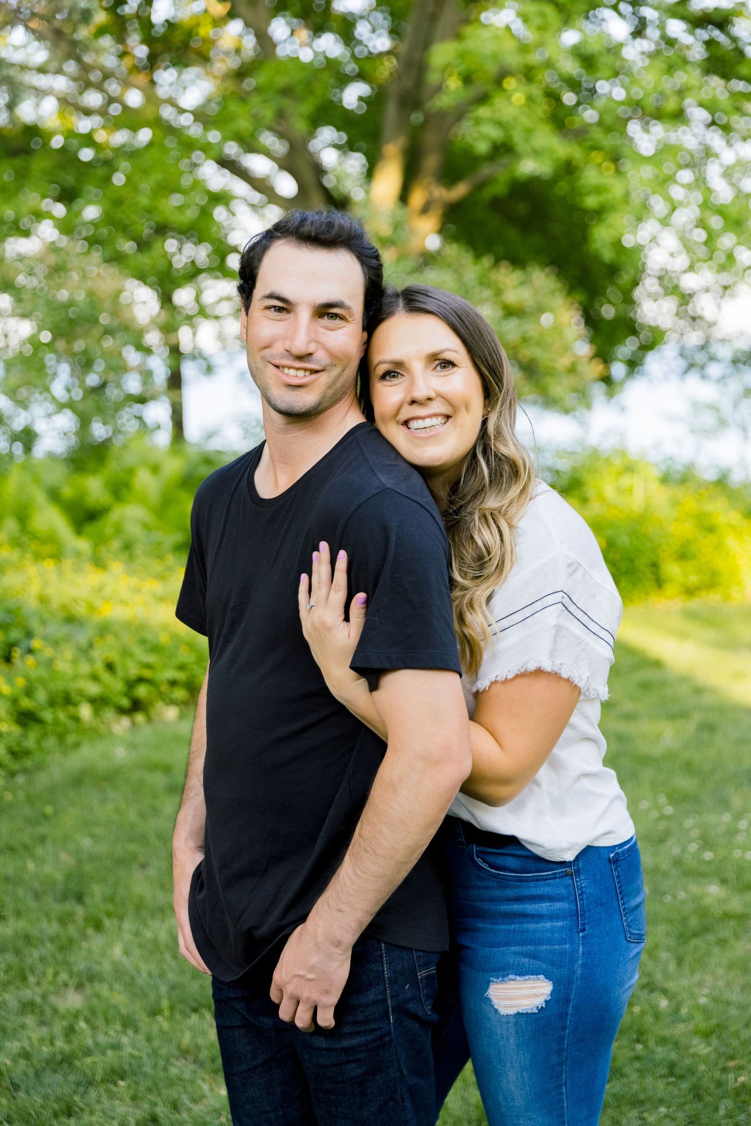 A couple in casual attire poses together outdoors with bright greenery in the background.