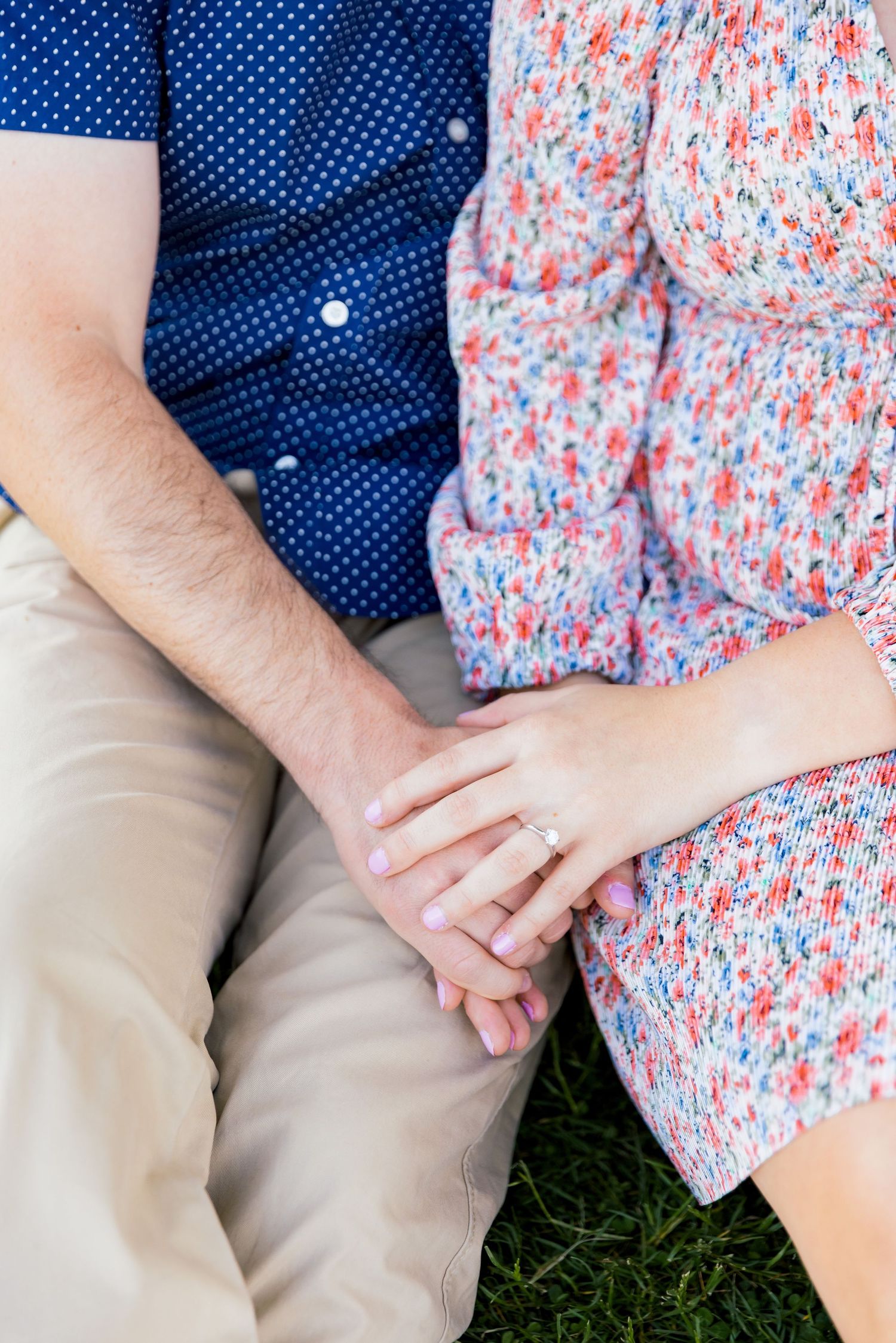 A couple holds hands while sitting on grass, wearing coordinating blue polka dot shirt and floral dress.