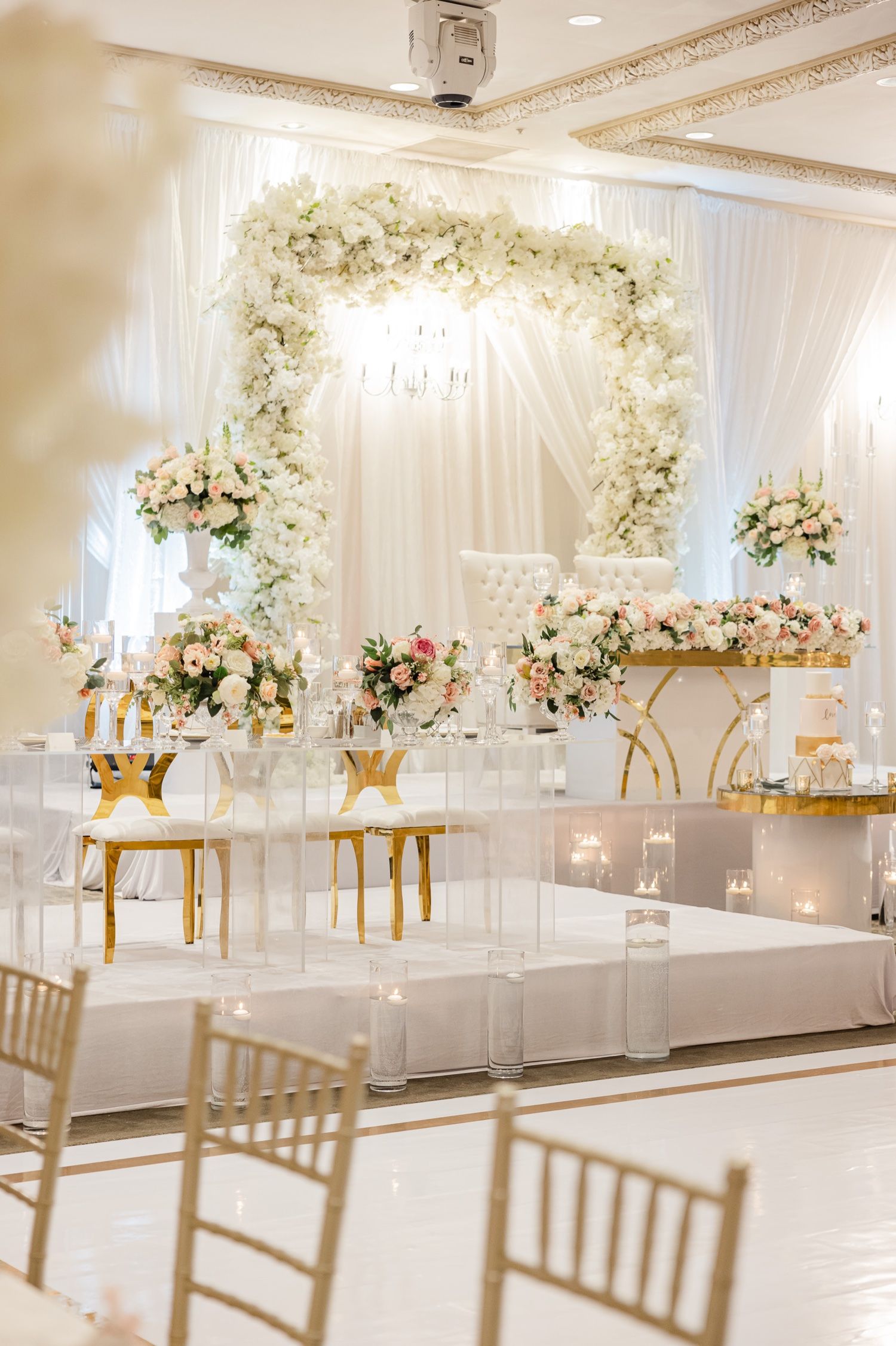 Elegant white floral arch wedding setup with gold accents and transparent chairs in a modern ballroom.