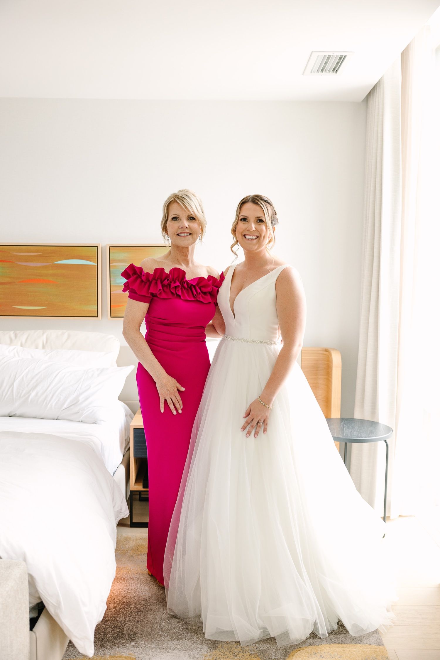 A woman in a hot pink ruffled gown poses with another in a white wedding dress in a modern hotel room.