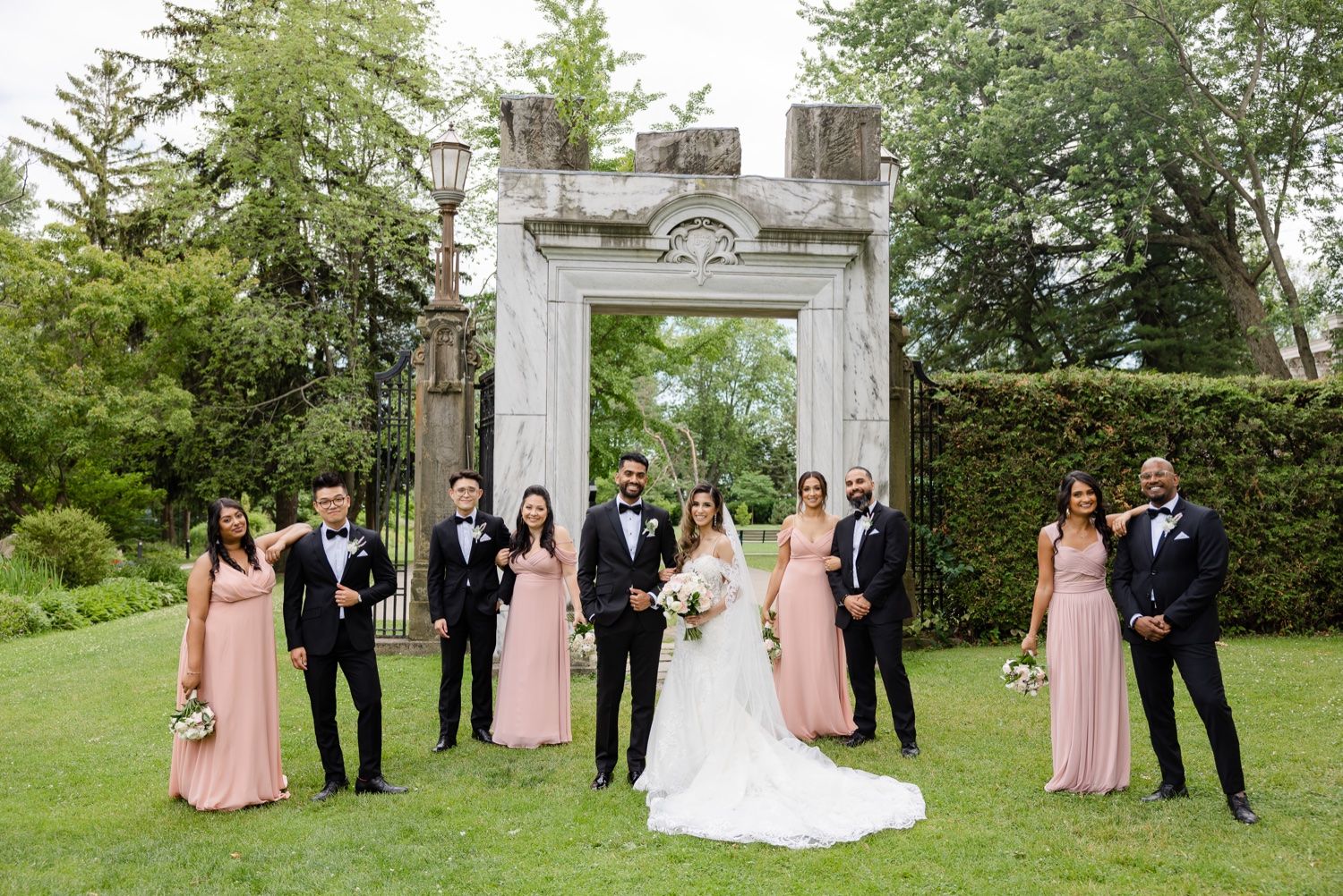 Wedding party arranged in front of a decorative stone gateway with pink bridesmaid dresses and black tuxedos.