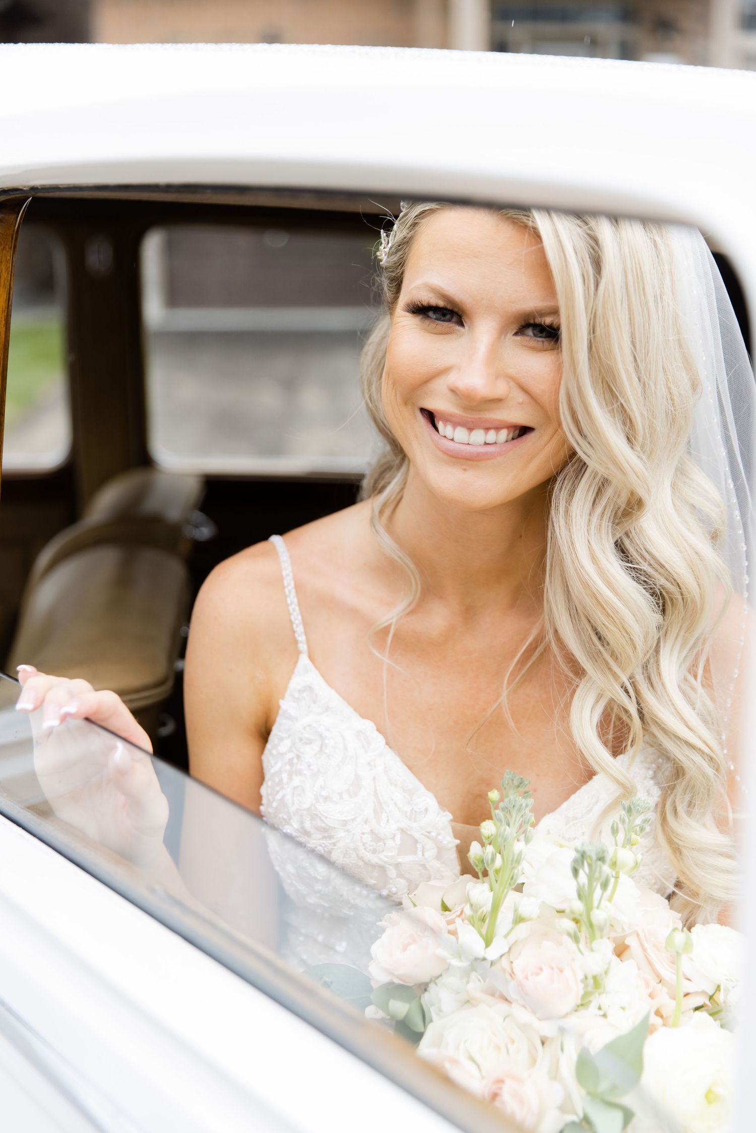 Bride with wavy blonde hair sits in vintage car while holding elegant floral bouquet.