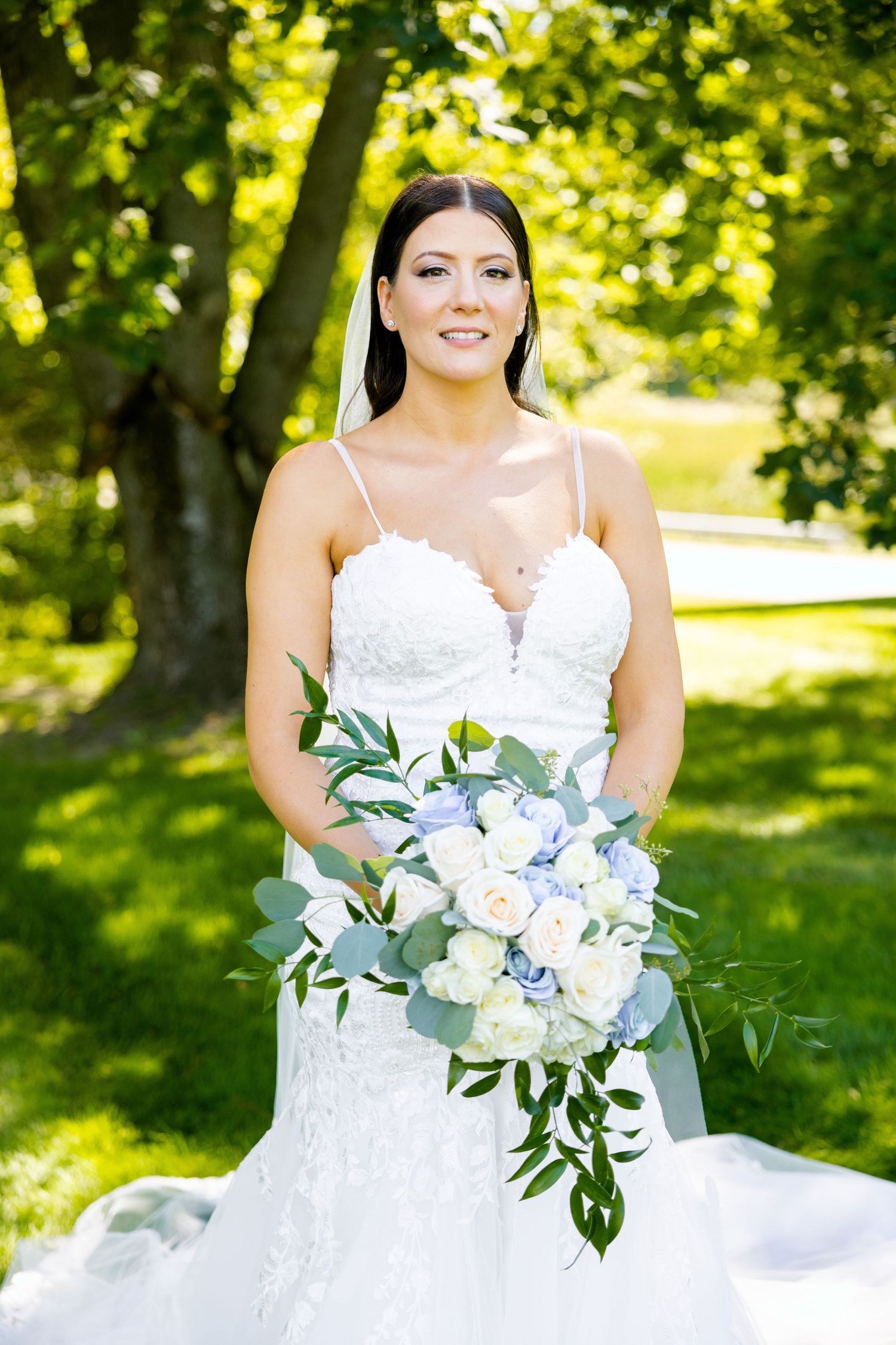 Bride in white lace gown holds elegant blue and white bouquet against lush green garden backdrop.