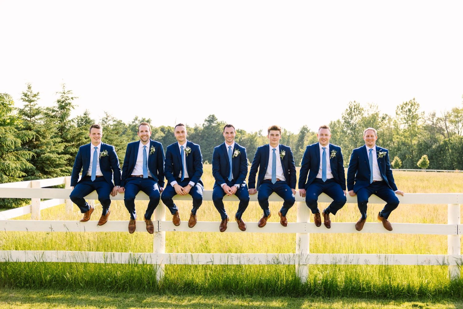 Groomsmen in matching navy suits sit together on white fence for fun wedding photo.