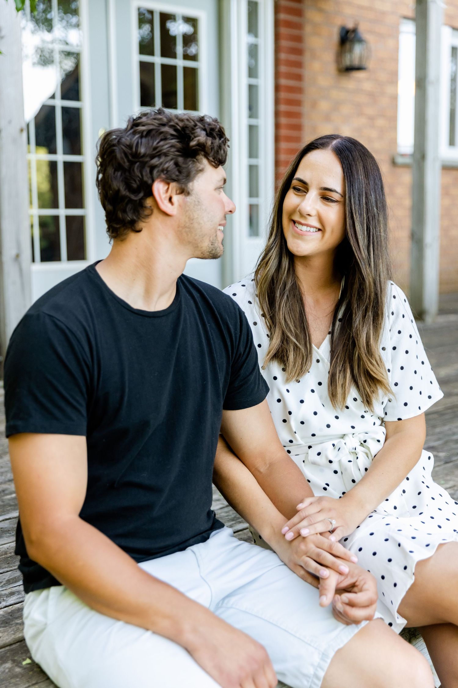 Couple sitting on porch steps, wearing black t-shirt and polka dot dress while sharing happy moment.