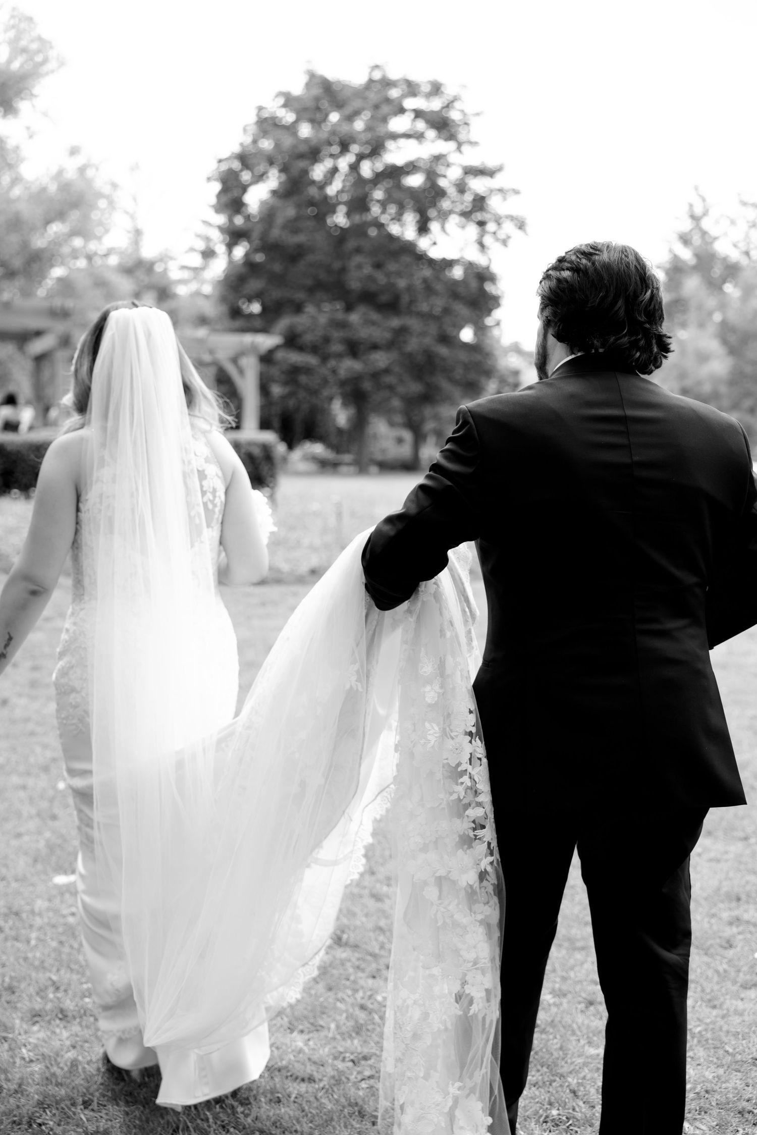 Black and white wedding photo of couple walking with flowing veil in outdoor setting.
