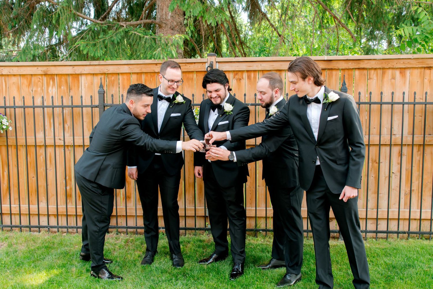 Group of groomsmen in tuxedos doing a celebratory fist bump in outdoor setting.
